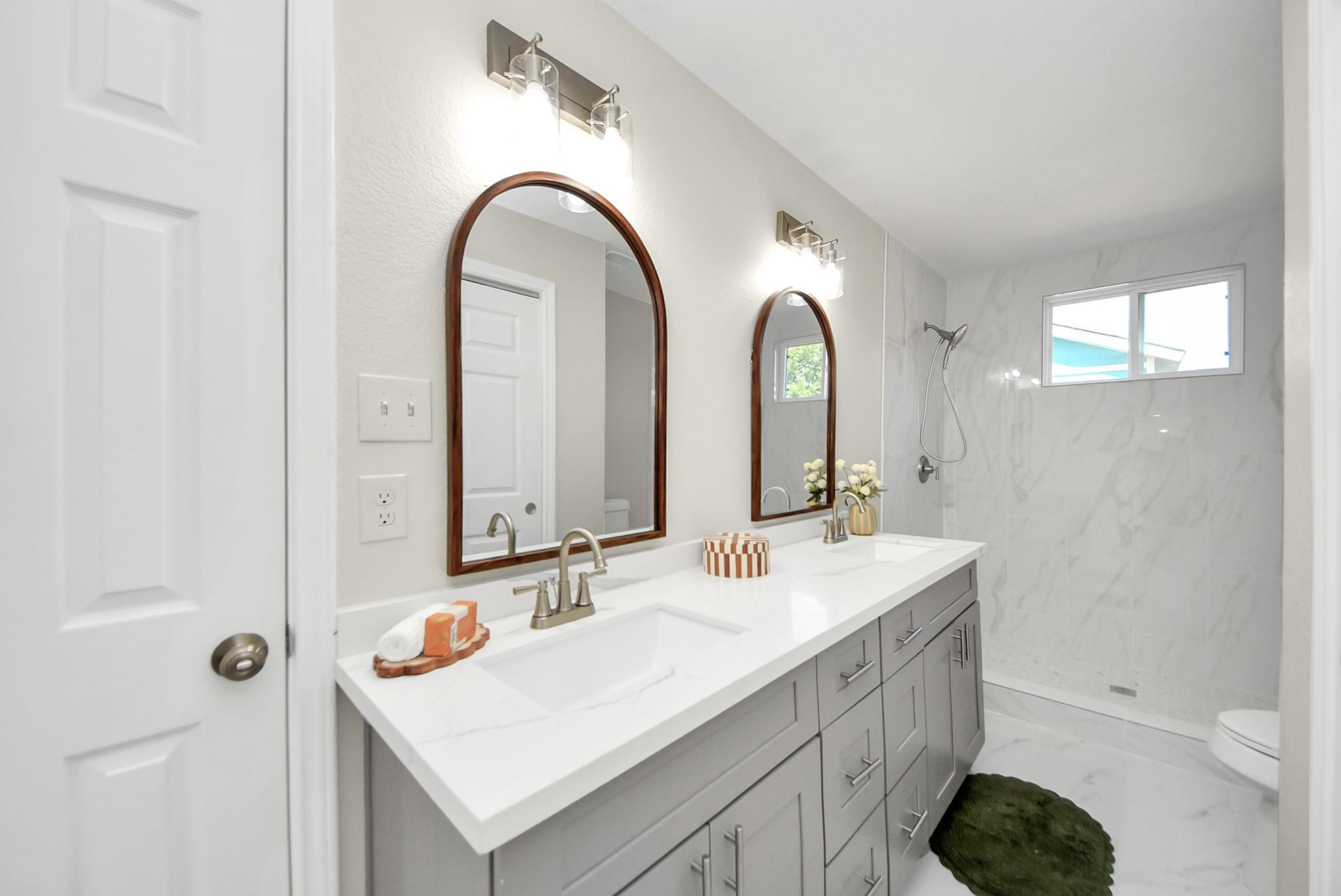 Bathroom with a gray double vanity, arched wood-framed mirrors, and a marble-look walk-in shower.