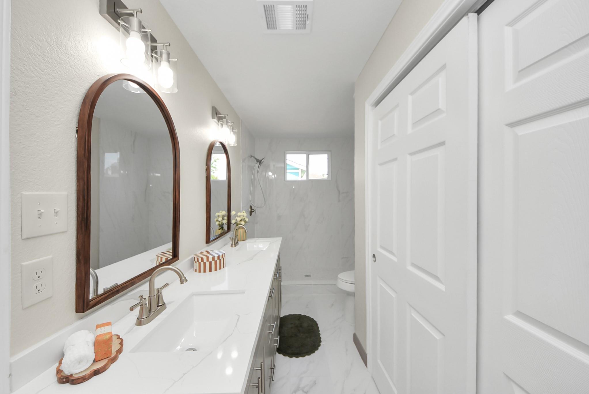 Bathroom with a long marble-look vanity, two arched wood-framed mirrors, and a tiled walk-in shower.