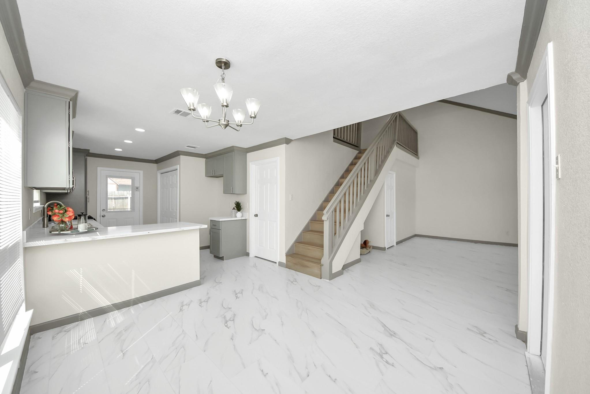 Open-plan kitchen and living area with a white peninsula, marble-look floors, and a staircase.