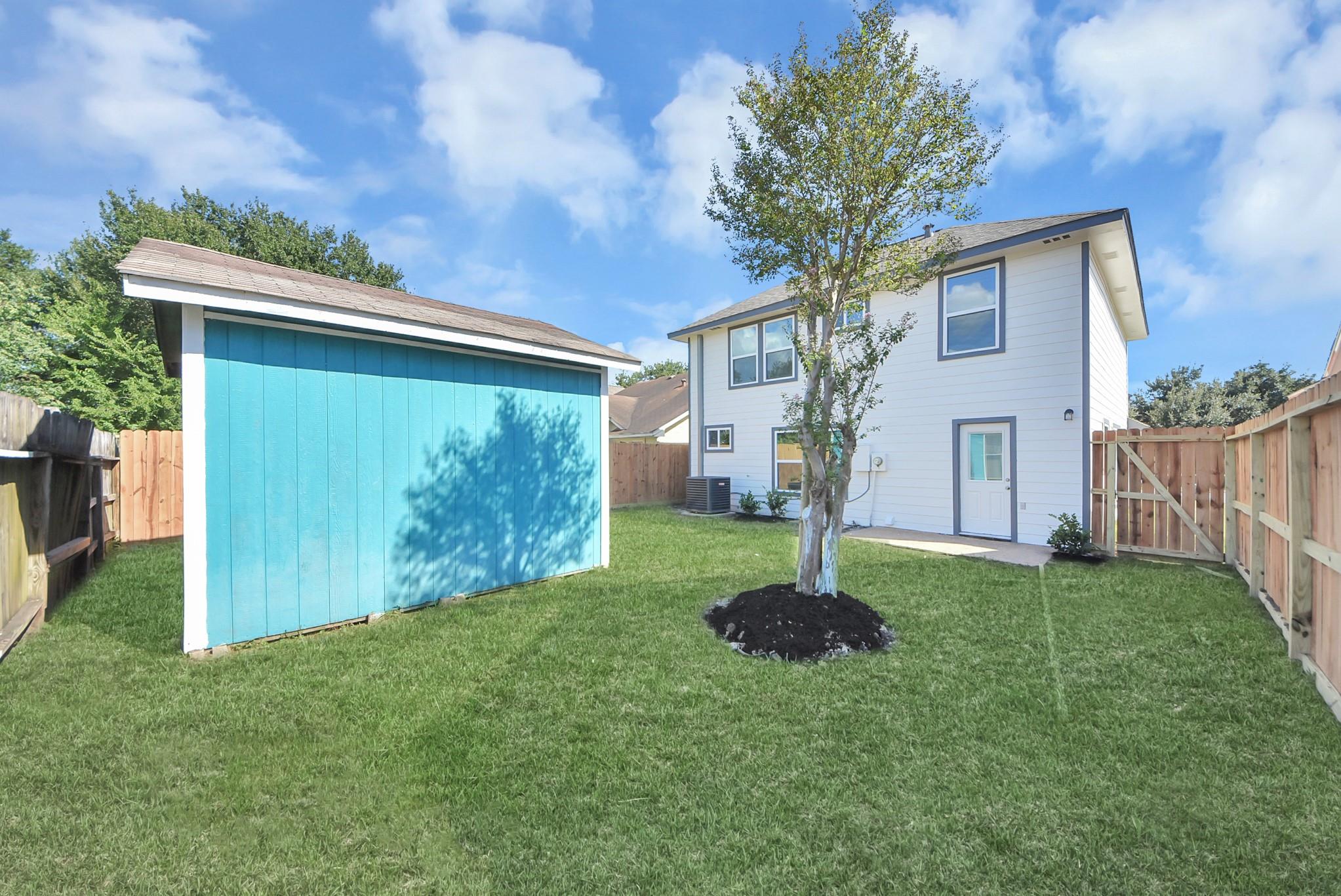Fenced backyard with a turquoise shed, grassy lawn, and leafy tree behind a two-story white house.