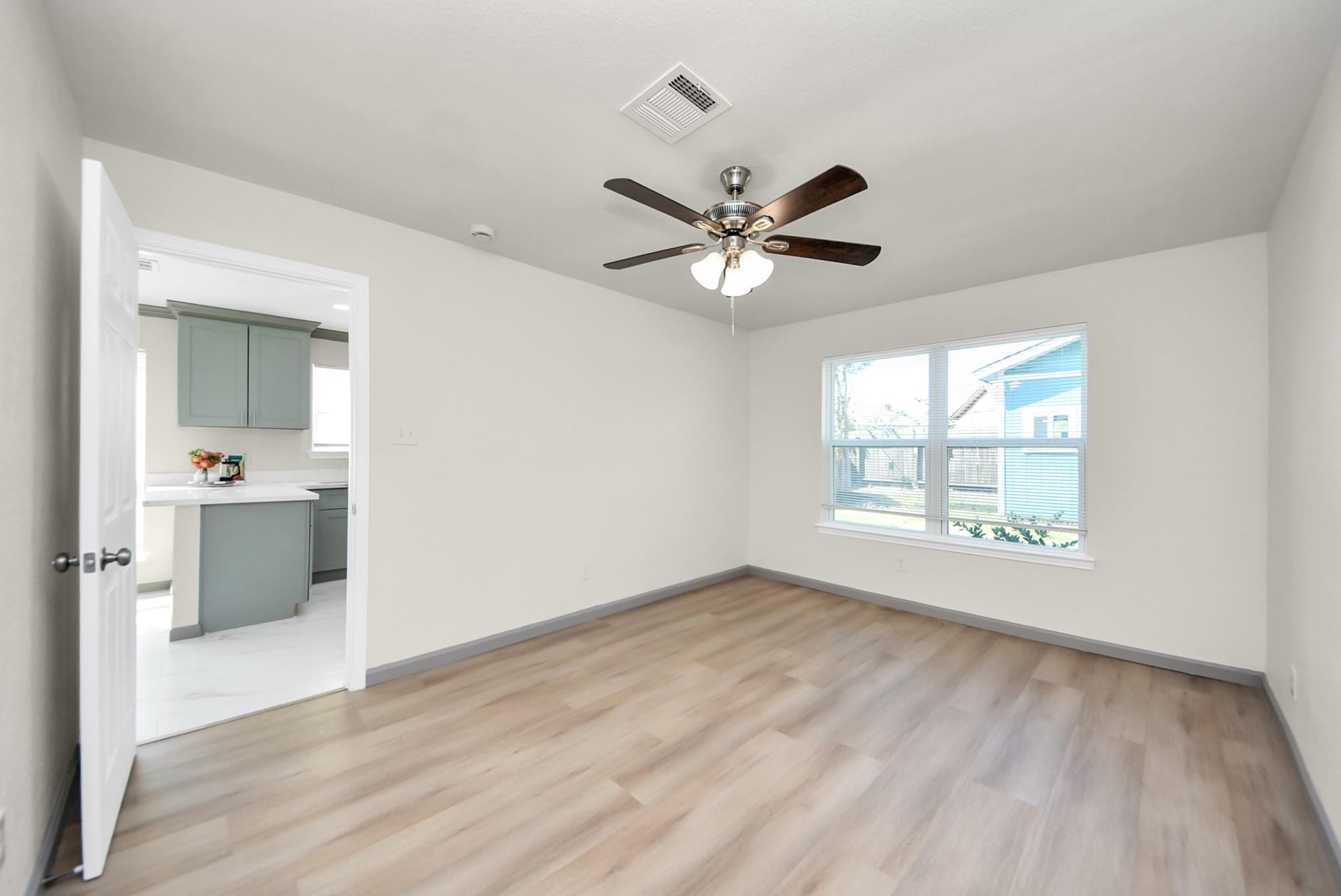 Empty room with wood-look floors, ceiling fan, and a window; kitchen visible through the doorway.