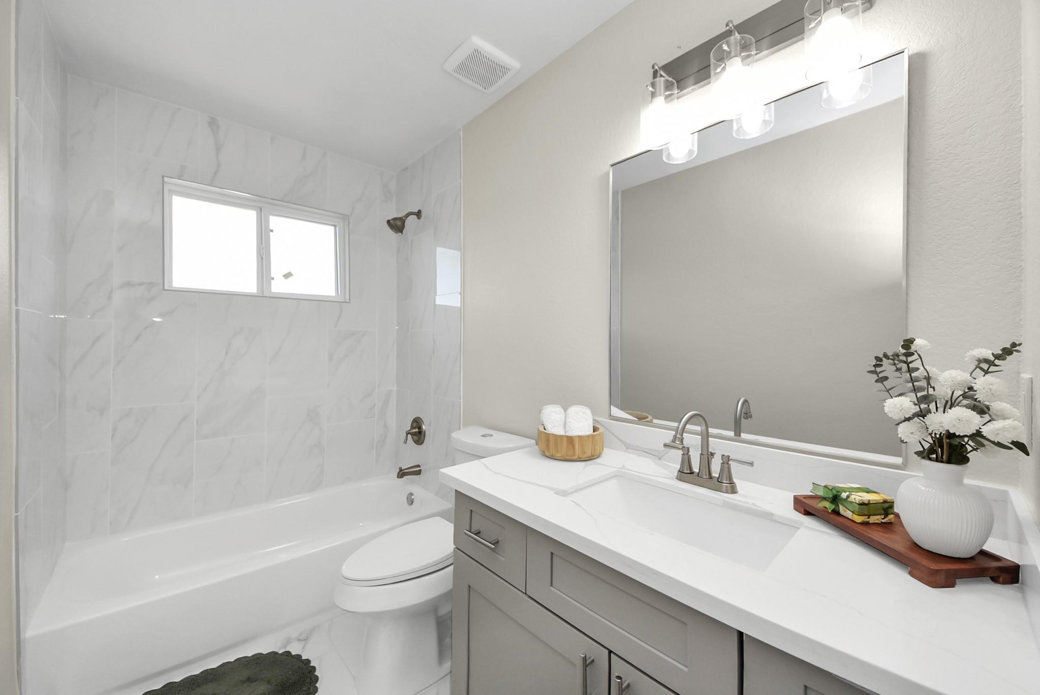 Bathroom with a marble-look tub surround and gray vanity topped by a large mirror.