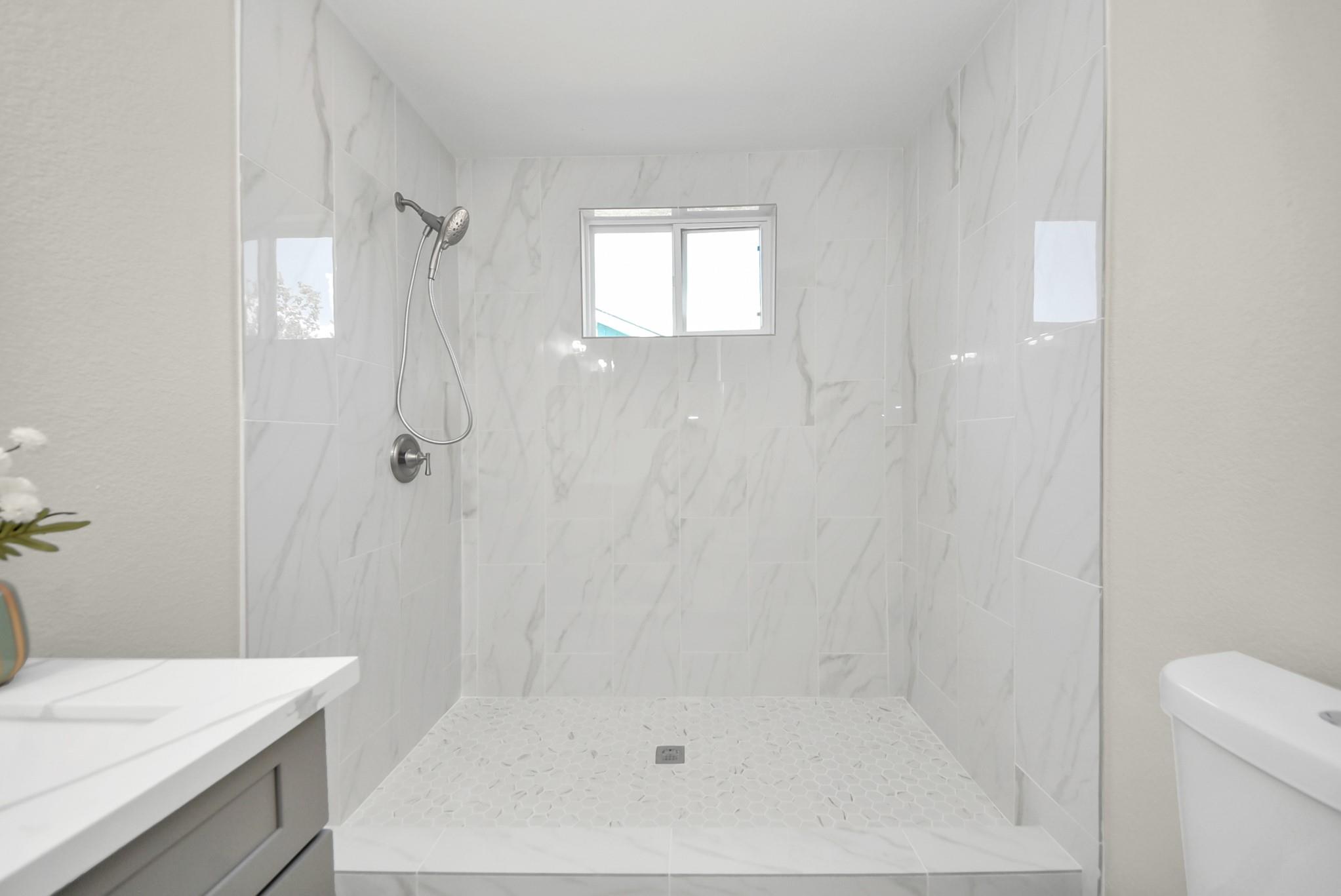 Walk-in shower with white marble-look tile, hexagonal floor, handheld showerhead, and high window.