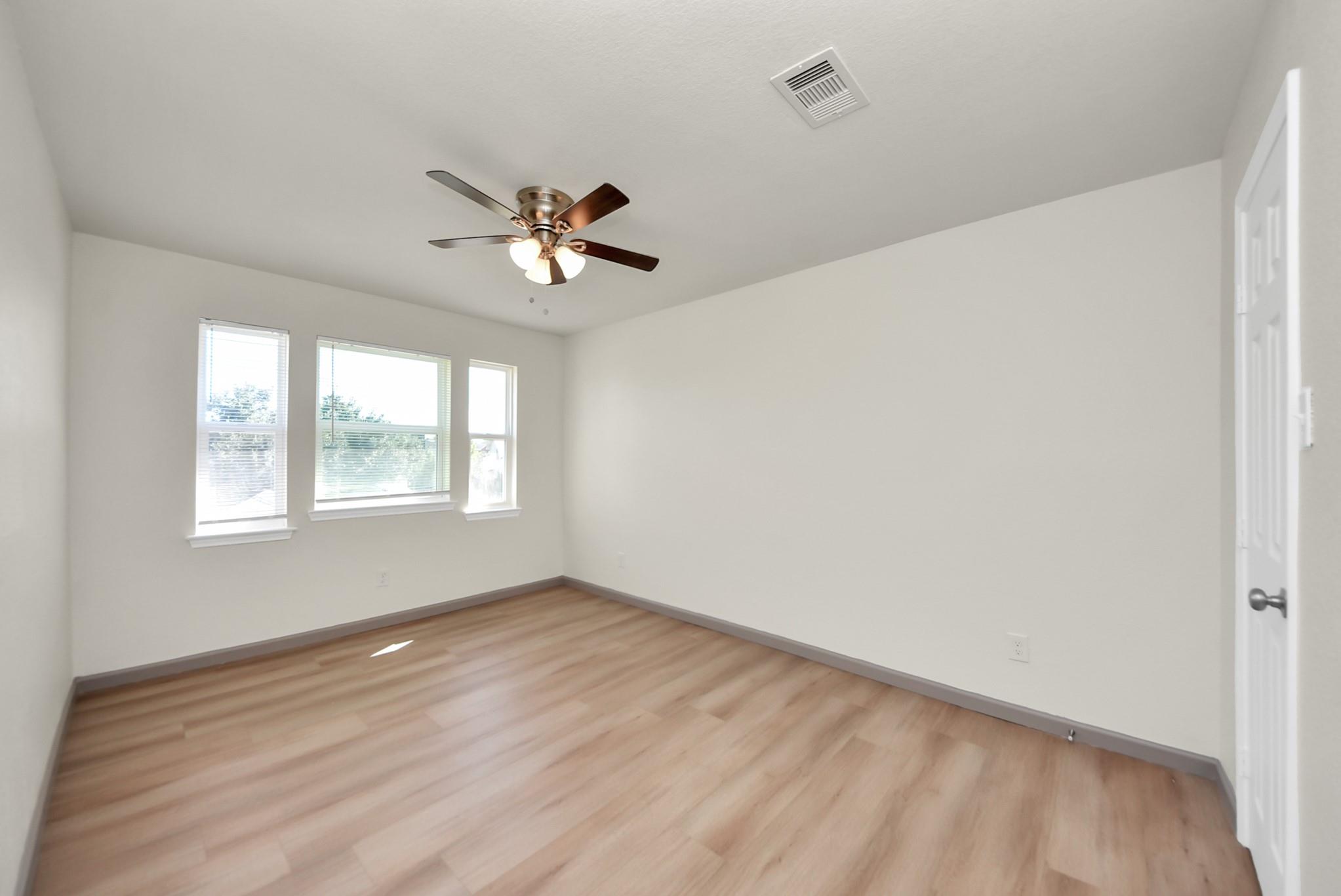 Empty room with light wood floors, three windows, and a ceiling fan with lights.