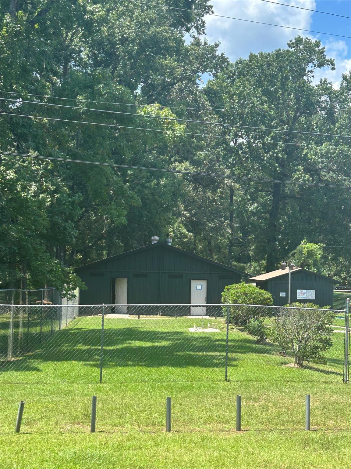 Two dark green buildings behind a chain-link fence, surrounded by lawn and mature trees.