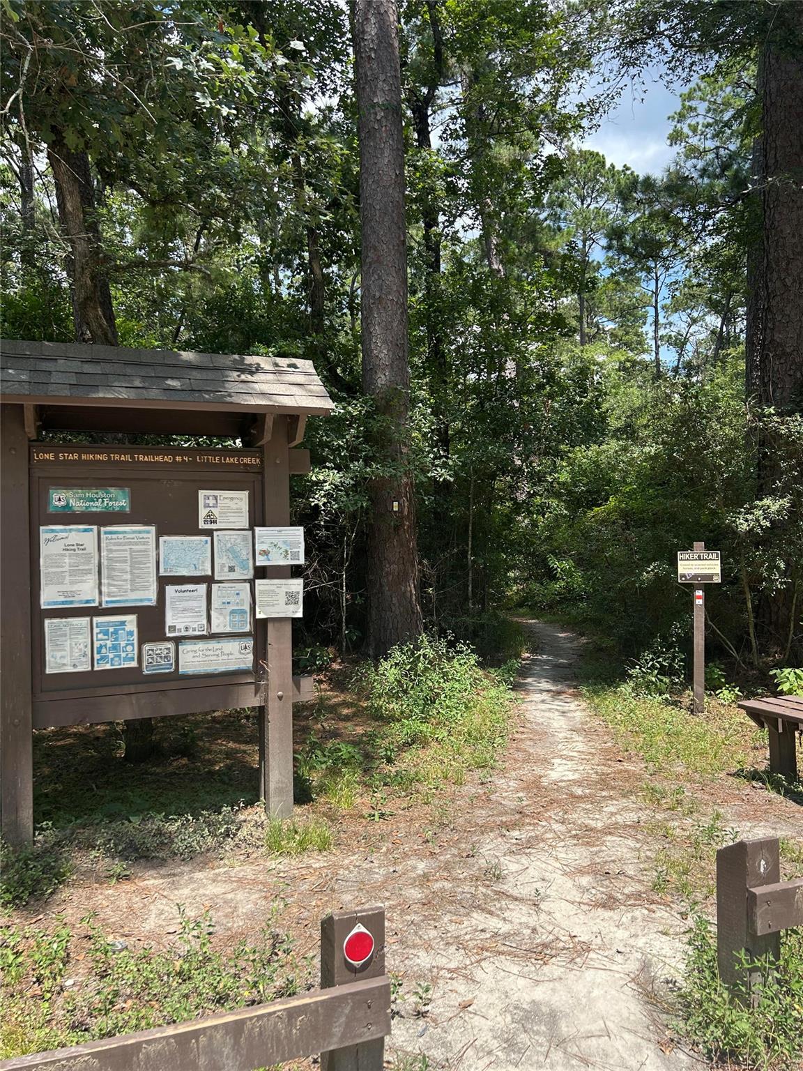 Wooded hiking trail entrance with an information board and a sandy path into the forest.