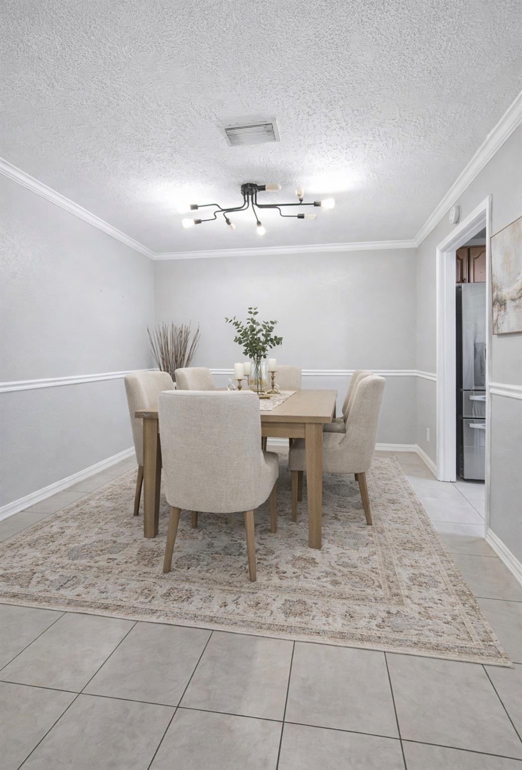 Staged dining room with a light wood table, upholstered chairs, patterned rug, and modern ceiling light.