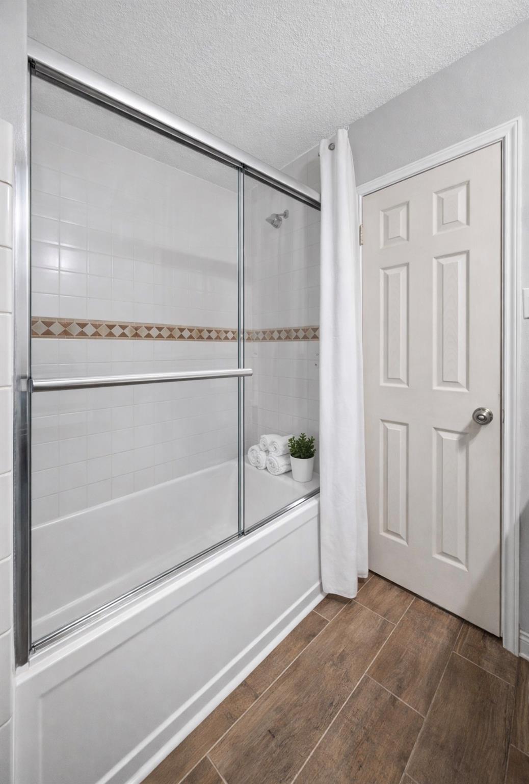 Bathroom with a glass-enclosed white tub, decorative tile border, and wood-look flooring.