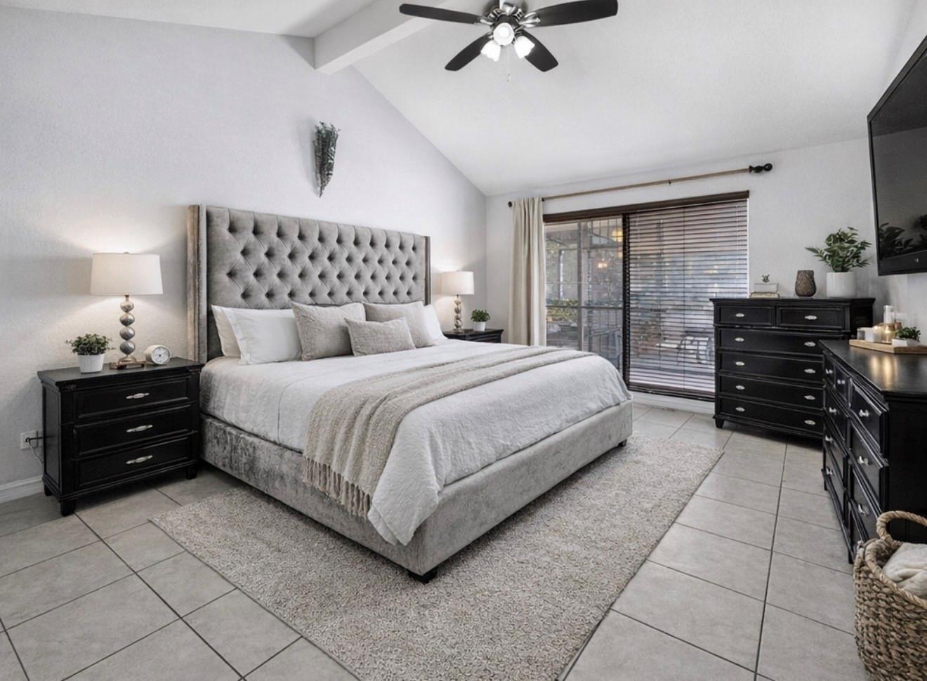 Bedroom with a tufted gray headboard, black dressers, and sliding glass doors with blinds.