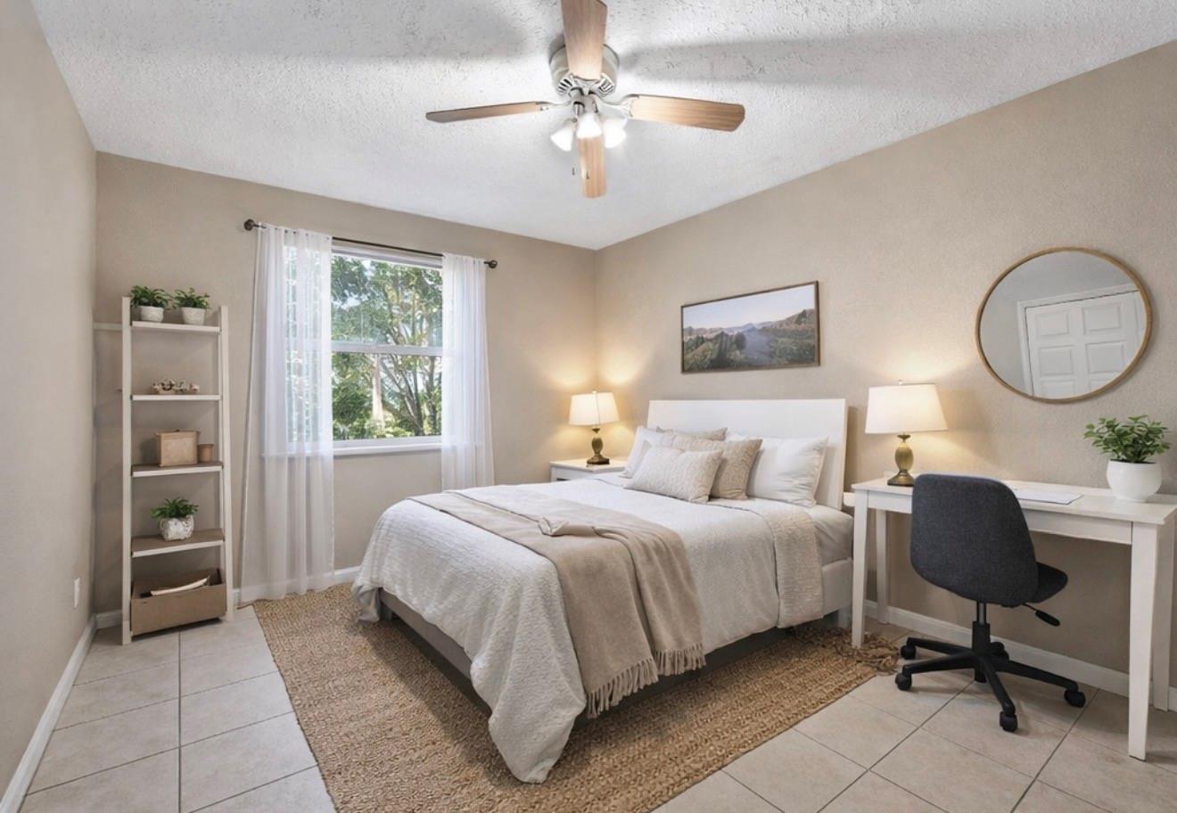 Furnished bedroom with a white bed, sheer-curtained window, ceiling fan, and desk.