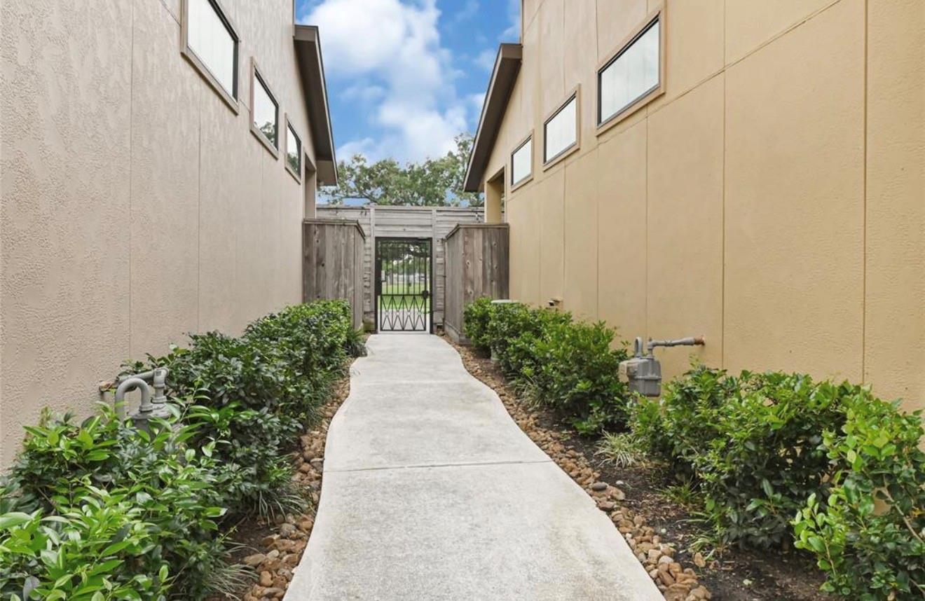 Concrete walkway between two buildings, bordered by shrubs and leading to a wooden gate.