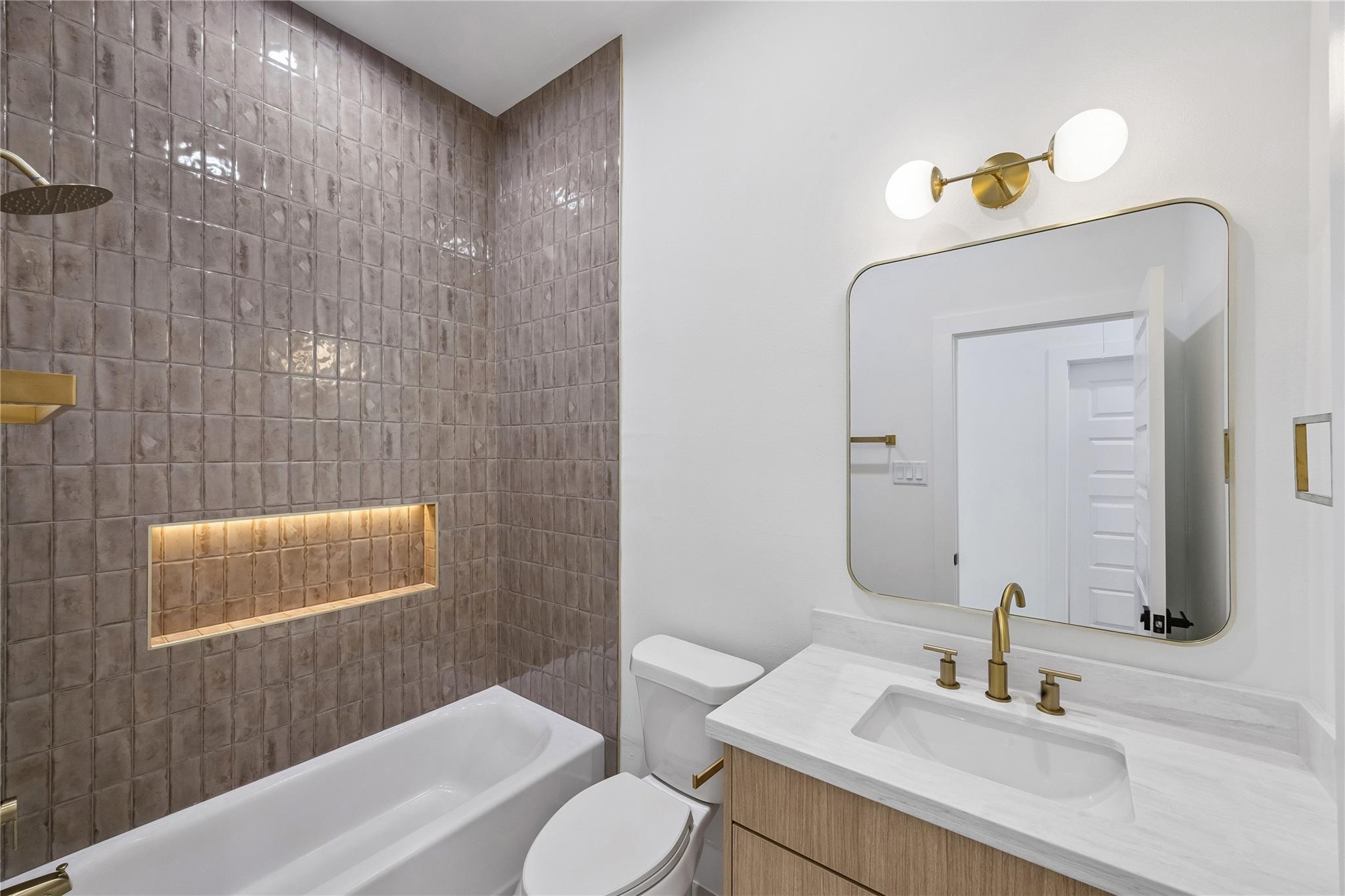 Bathroom with a tiled tub and shower, illuminated niche, brass fixtures, and a wood vanity.