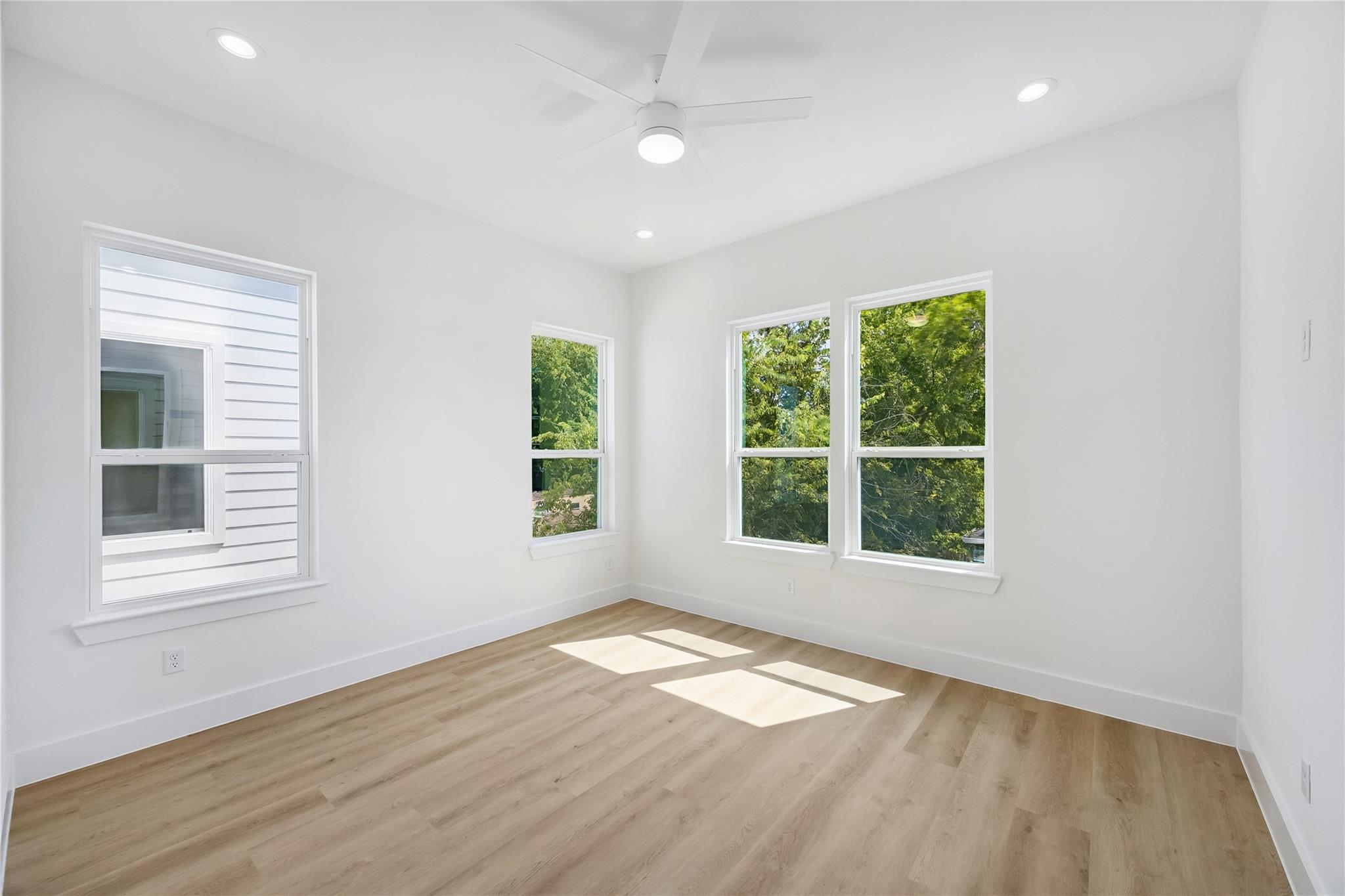 Empty room with wood-look flooring, a ceiling fan, and four windows overlooking trees.