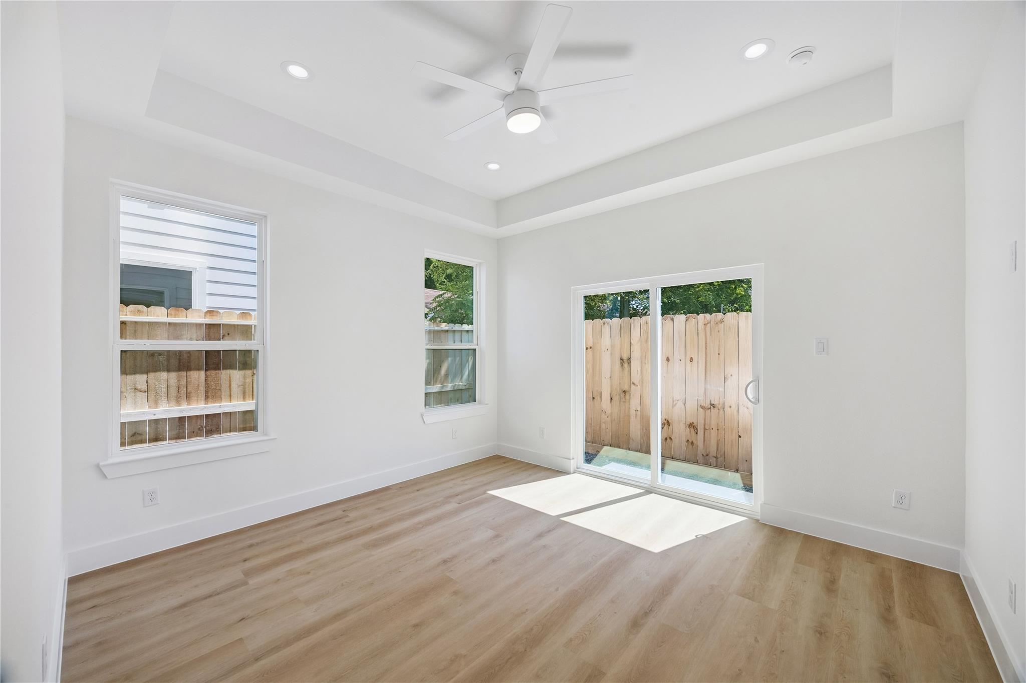 Bright, vacant room with wood-look floors, ceiling fan, and sliding glass door to a fenced patio.