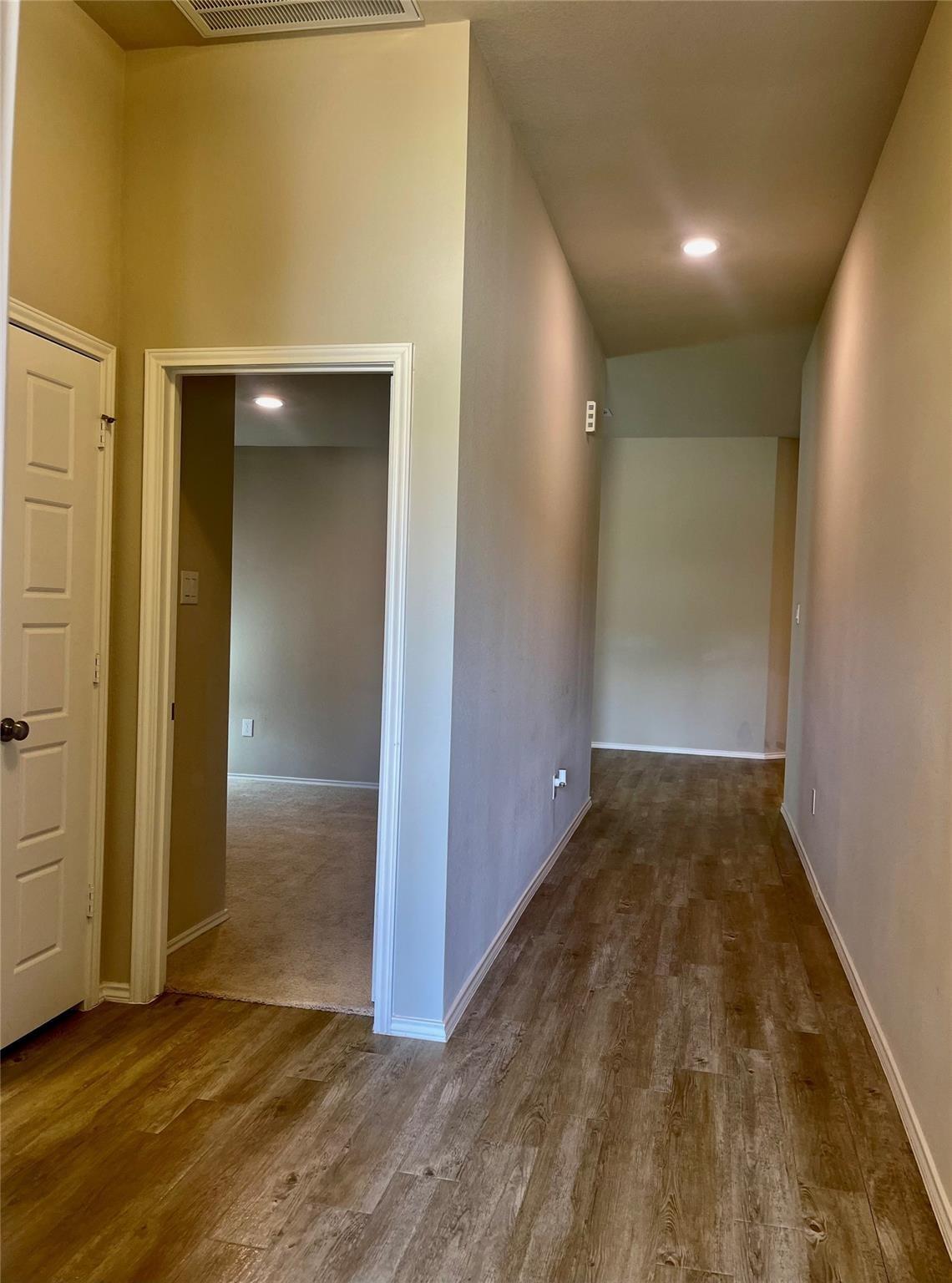 Empty hallway with wood-look plank flooring and an open doorway to a carpeted room.