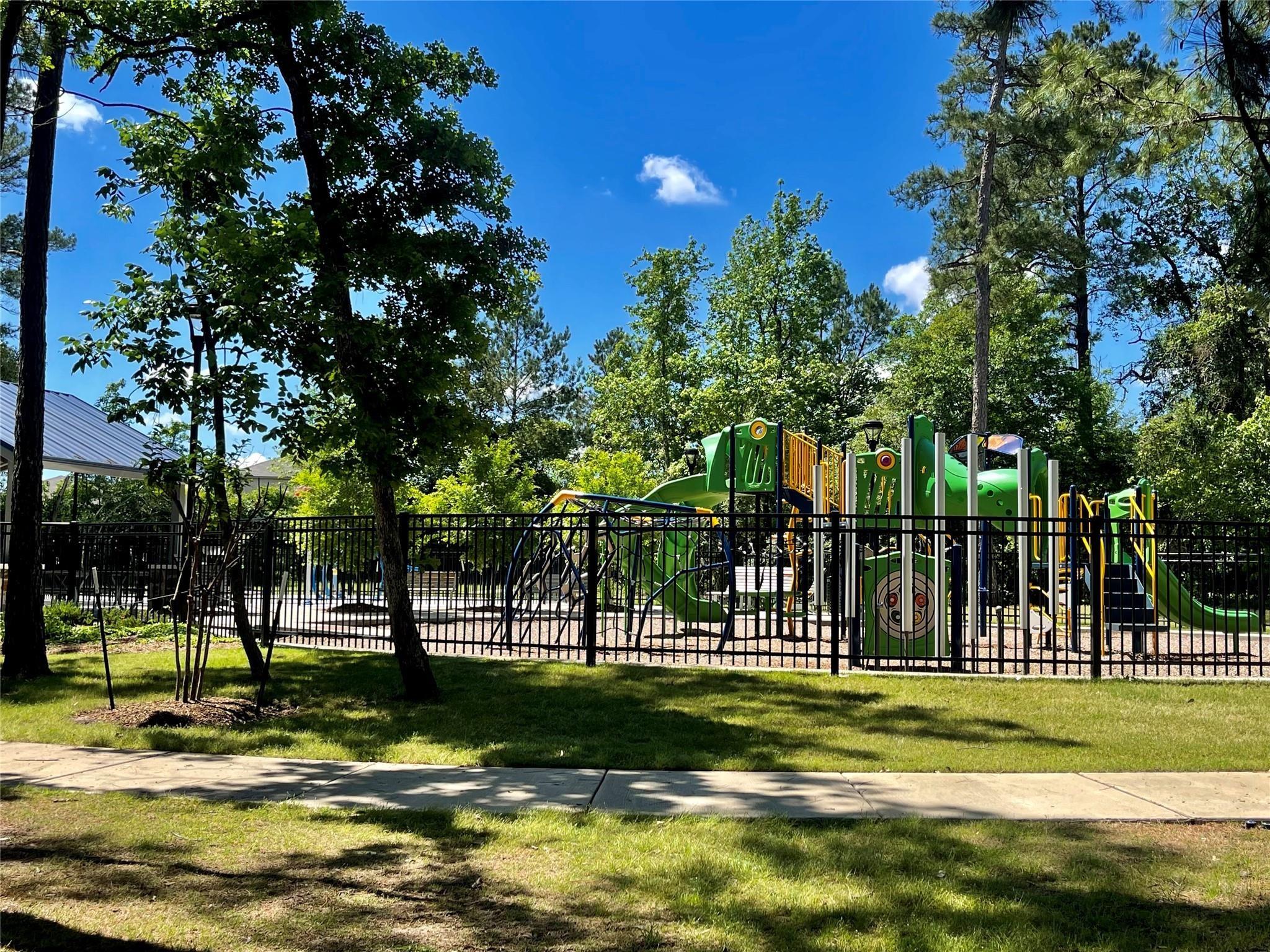 Fenced playground with colorful climbing structures and slides beneath mature trees.
