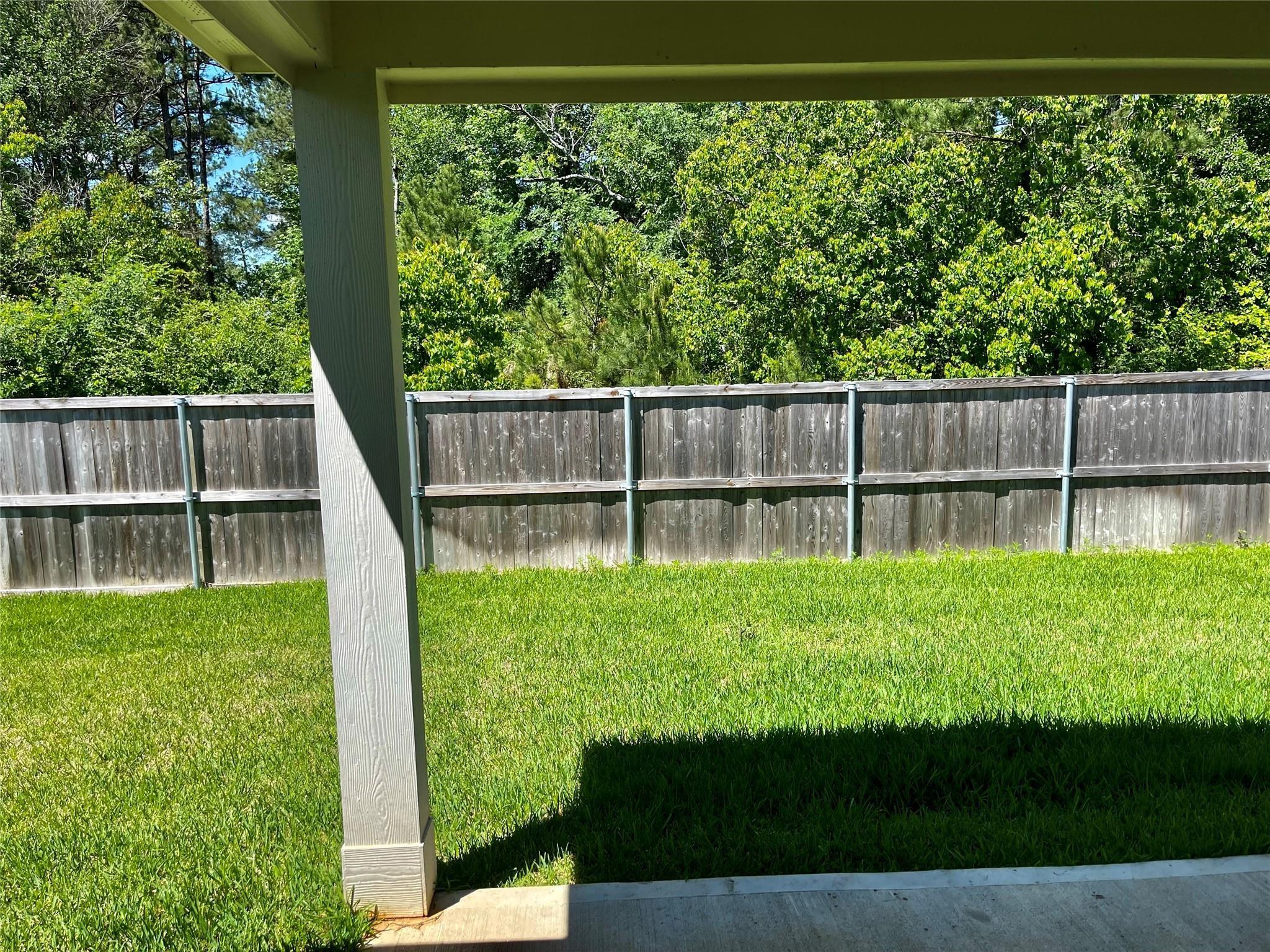 Covered patio overlooks a green lawn bordered by a wooden privacy fence and leafy trees.