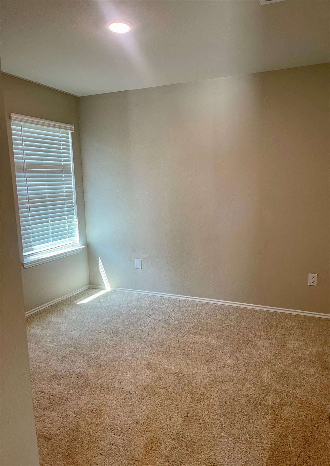 Empty carpeted room with beige walls, white window blinds, and sunlight on the floor.