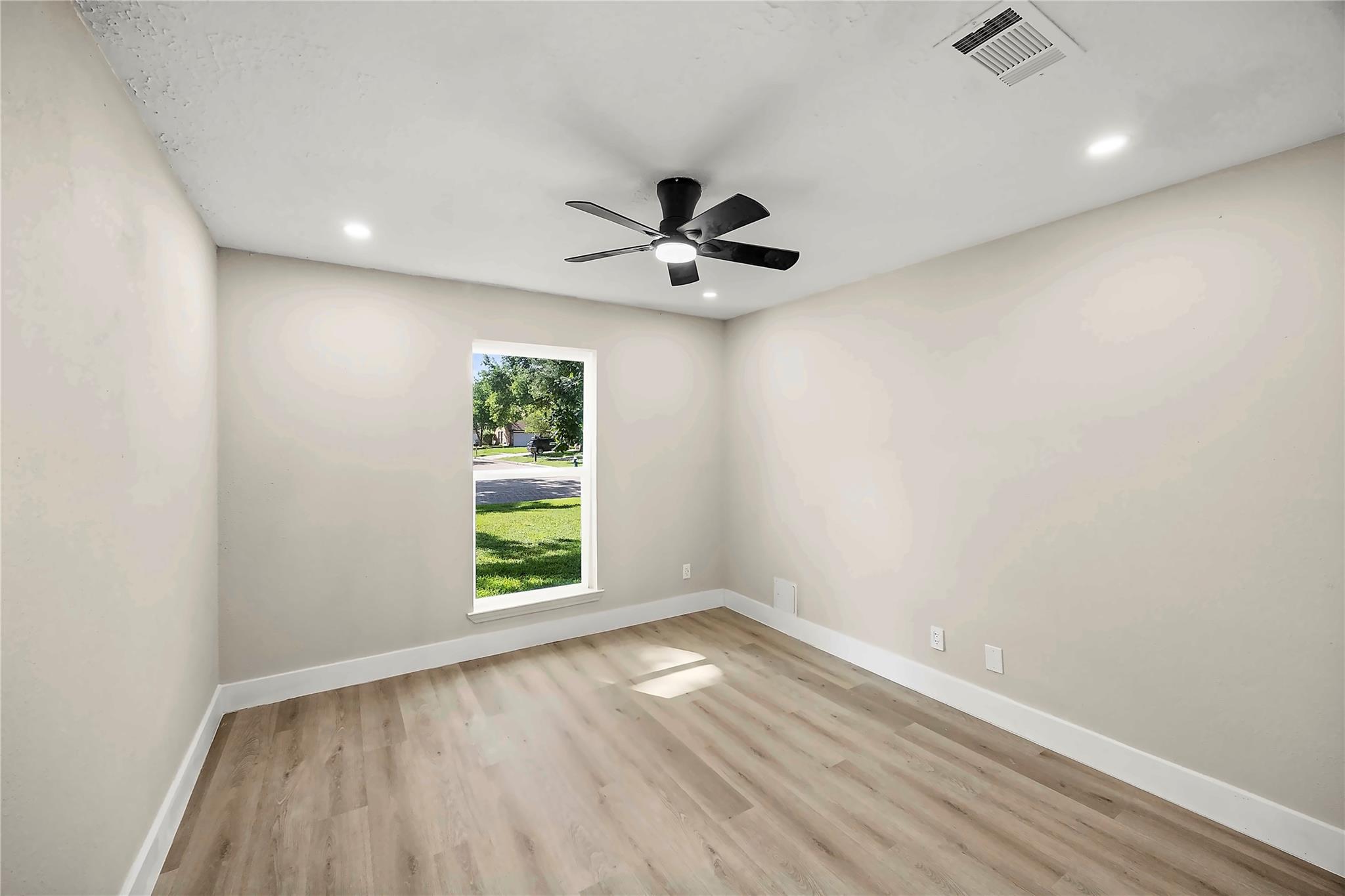 Empty bedroom with wood-look flooring, black ceiling fan, and a tall window overlooking a lawn.