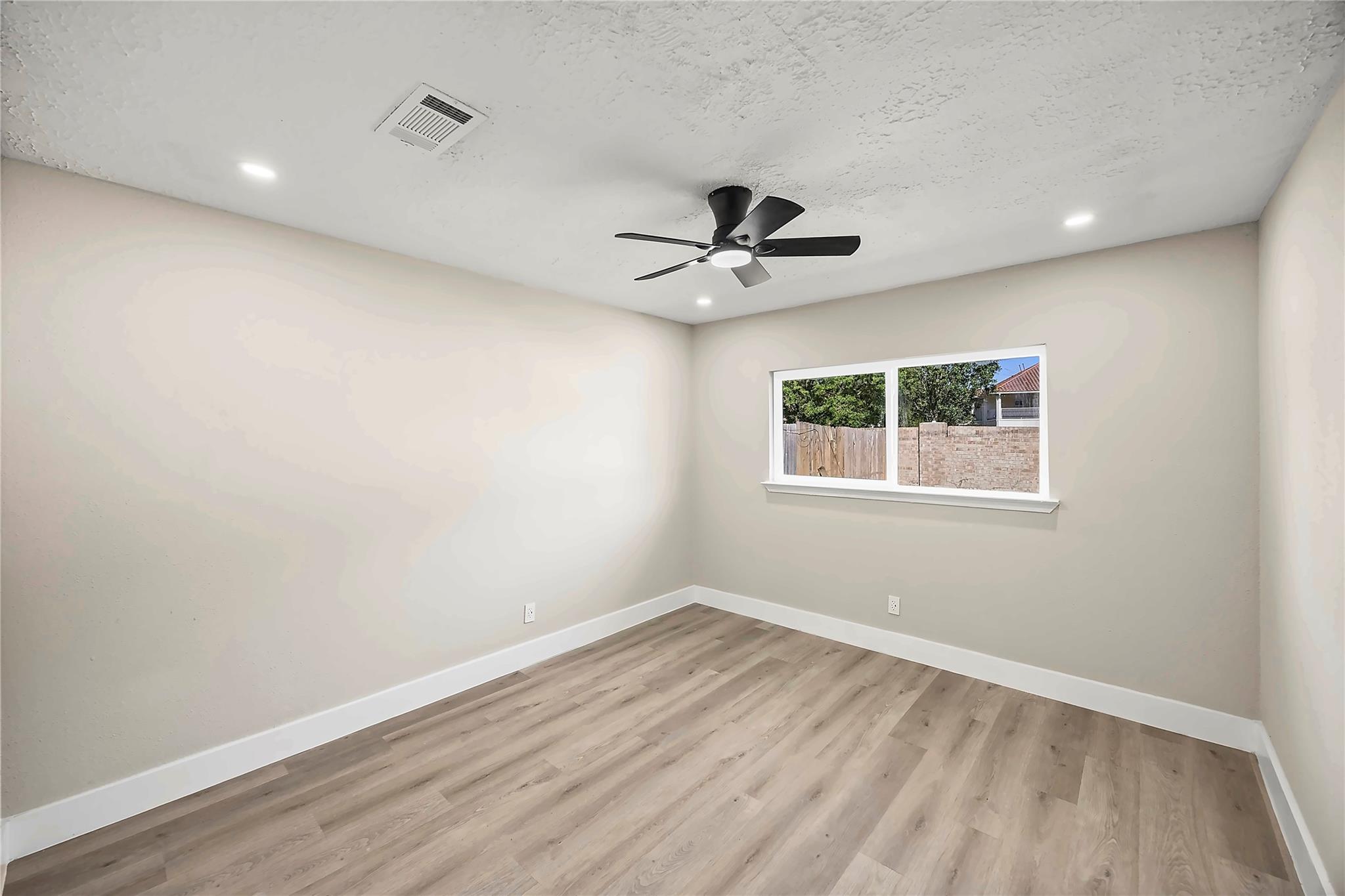 Empty bedroom with light wood-look flooring, a black ceiling fan, and a window overlooking a fenced yard.