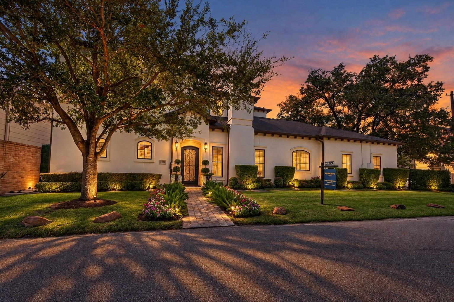 White stucco home with illuminated windows, landscaped entry path, and a sunset sky.