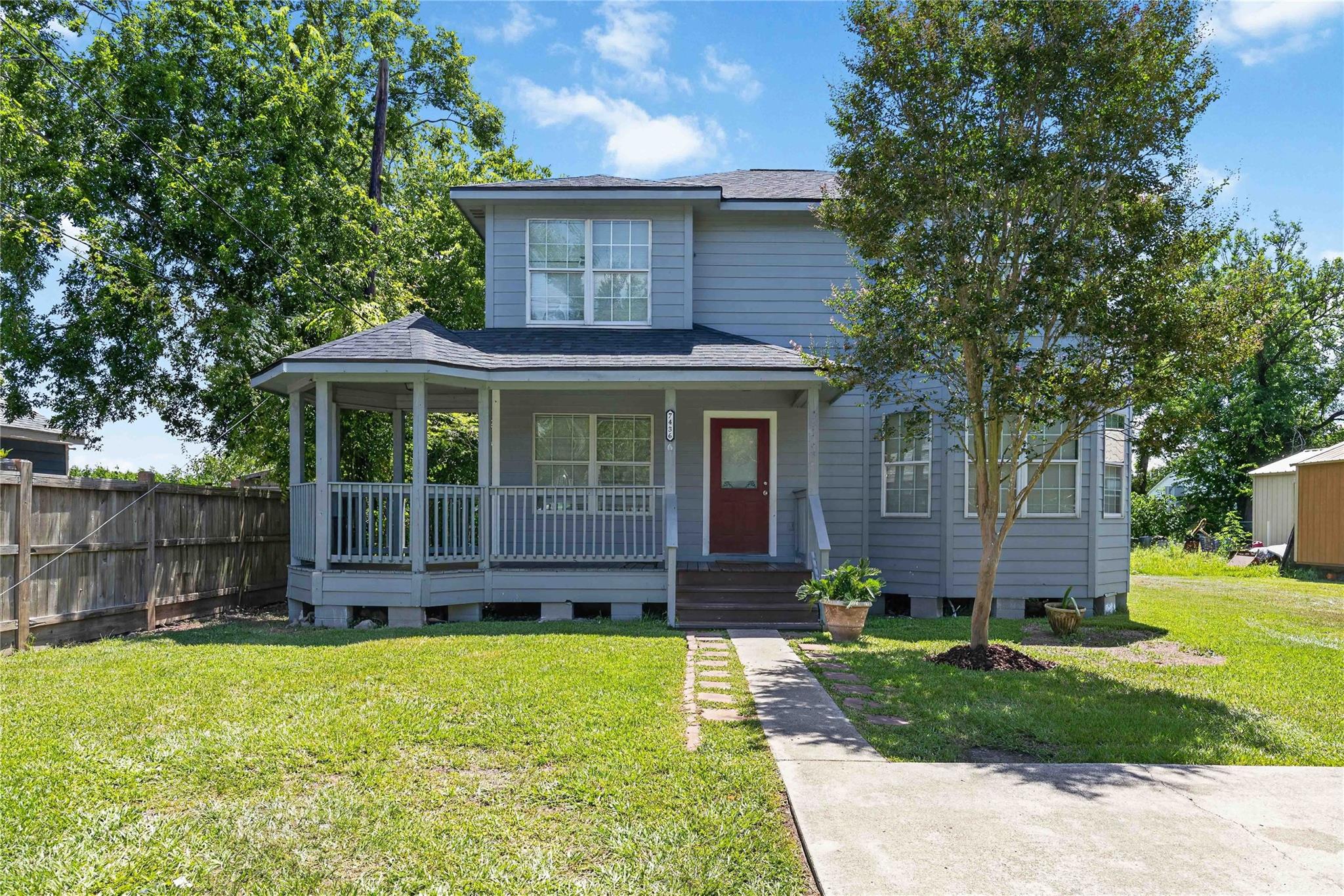 Two-story gray house with a covered porch, red front door, and mature tree in the front lawn.