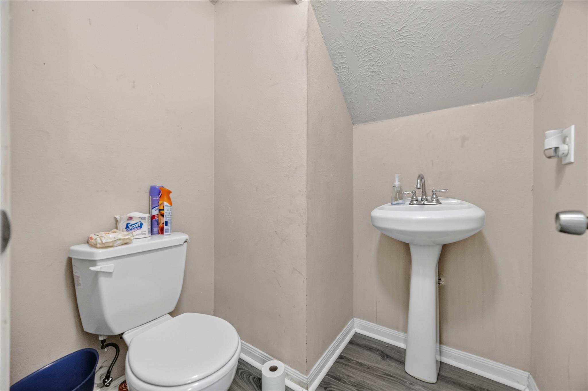 Bathroom with a white toilet and pedestal sink, beige walls, and gray wood-look flooring.