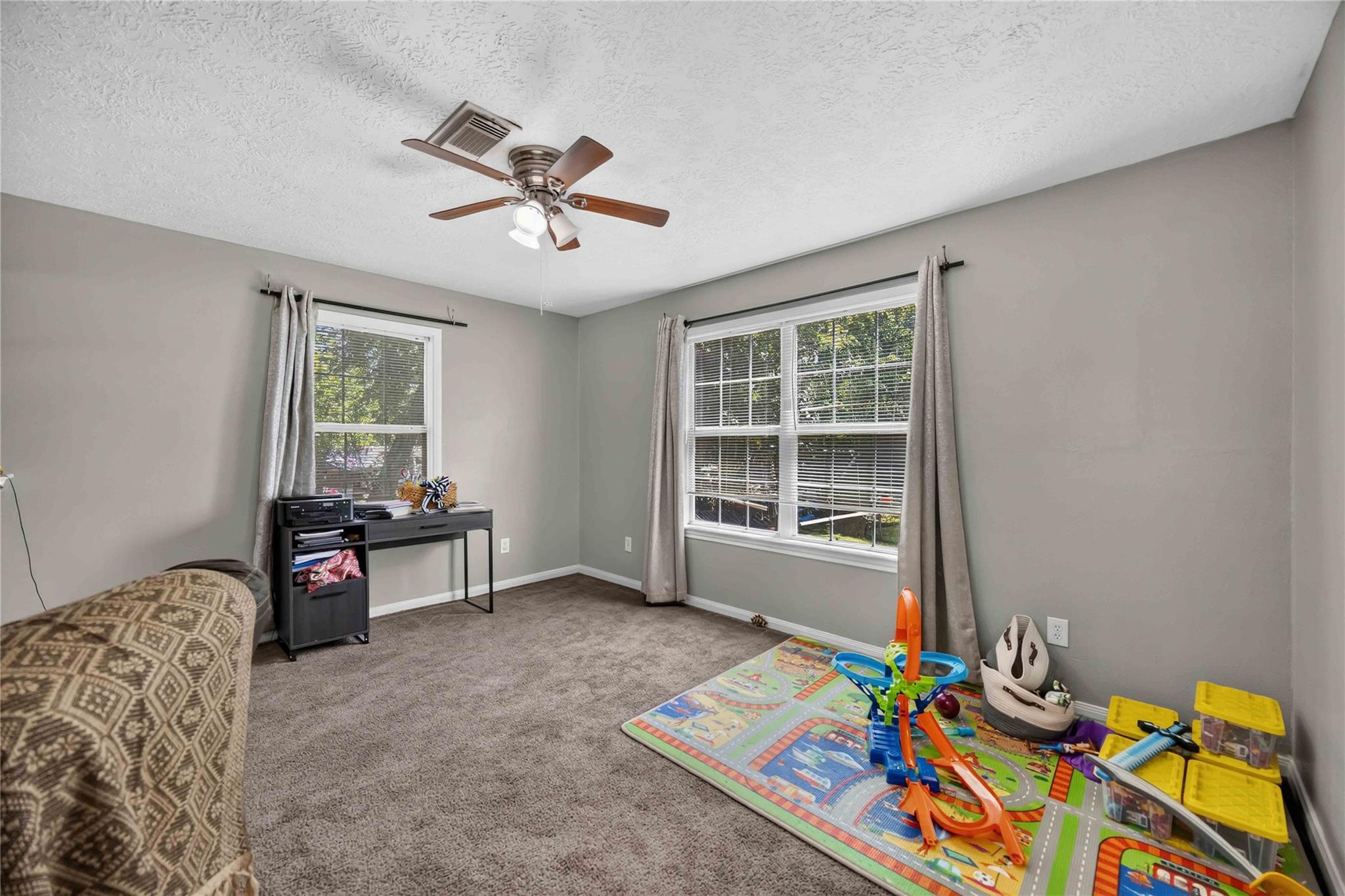 Carpeted playroom with two windows, a desk, and a colorful toy car track.
