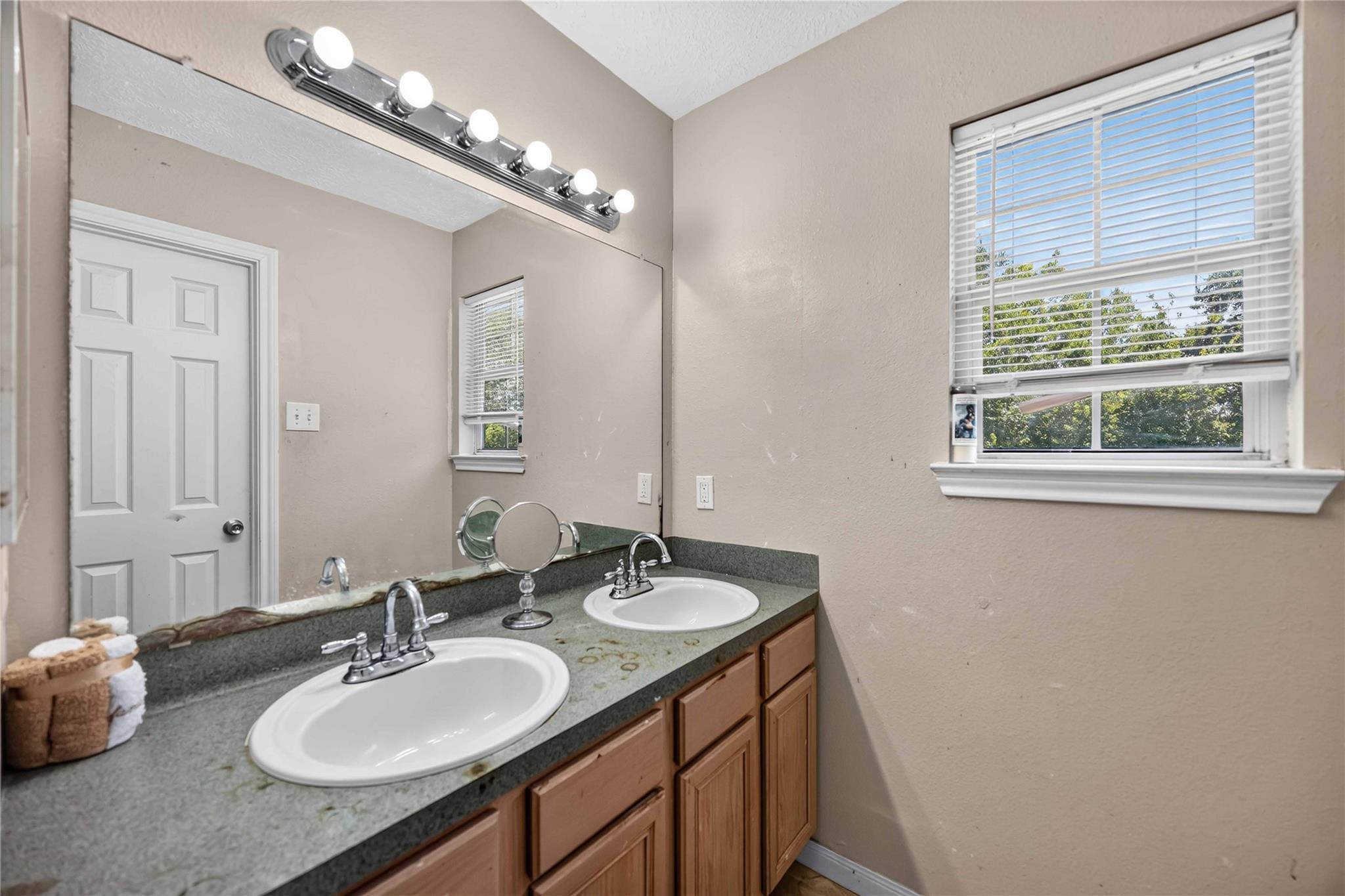 Bathroom double vanity with green countertop, wood cabinets, and a large mirror beside a window.