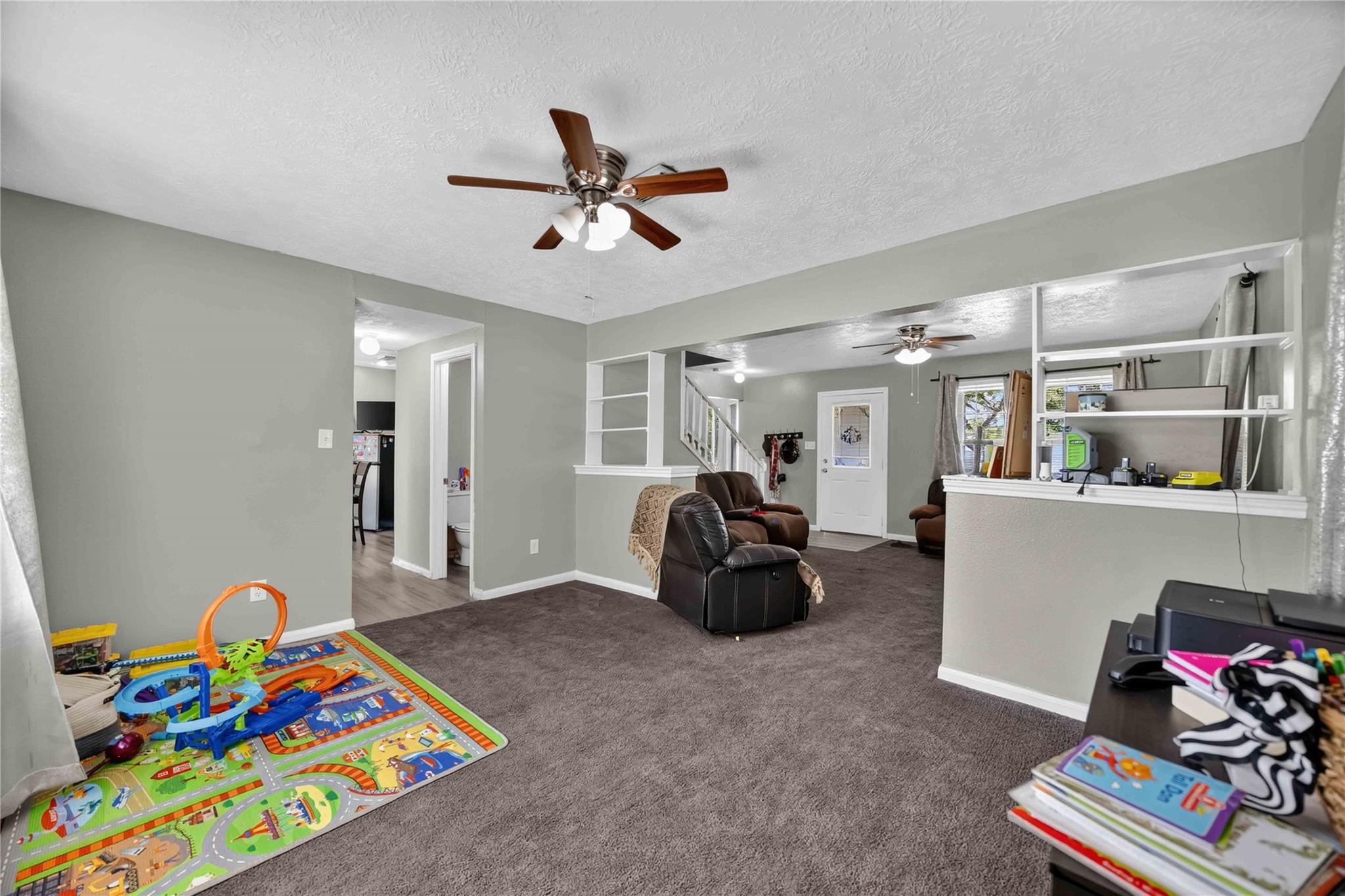 Furnished living room with gray carpet, leather recliners, ceiling fans, and a children's play area.