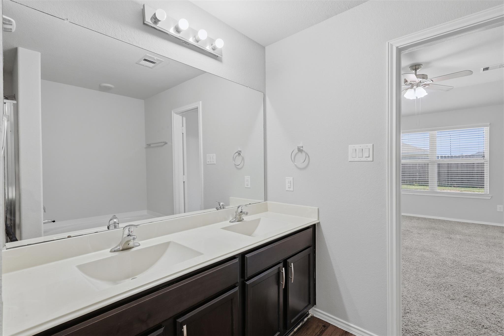 Bathroom double vanity with dark cabinets and a large mirror, beside an open doorway to a carpeted bedroom.