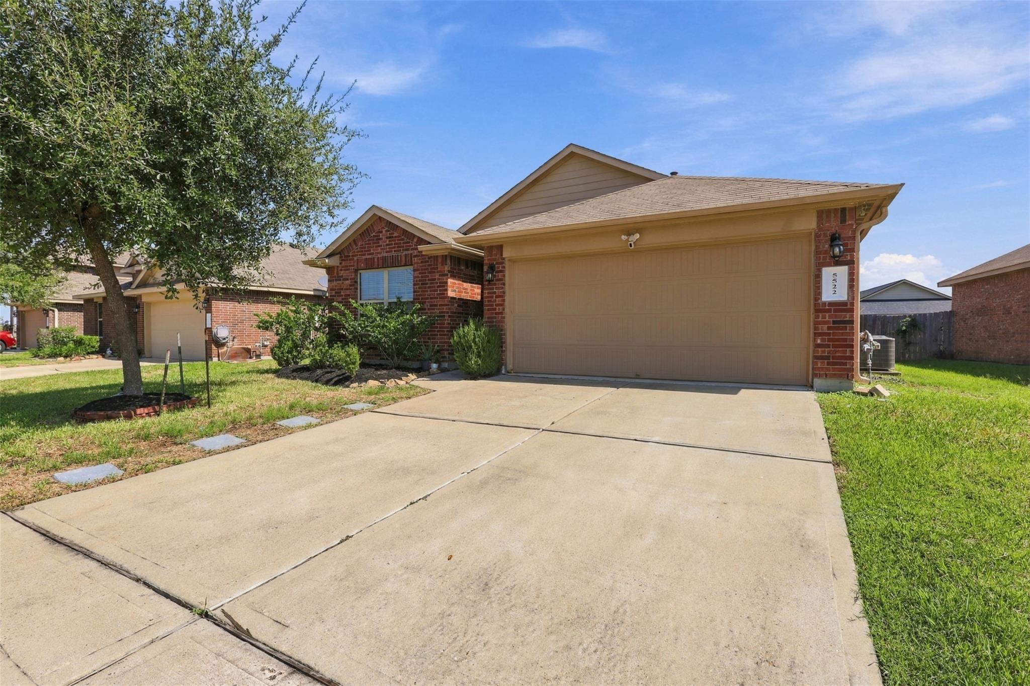 Red brick home with a tan two-car garage, broad concrete driveway, and mature front-yard tree.