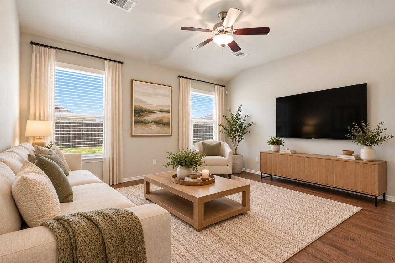 Staged living room with a cream sectional, wood furnishings, wall-mounted television, and two bright windows.