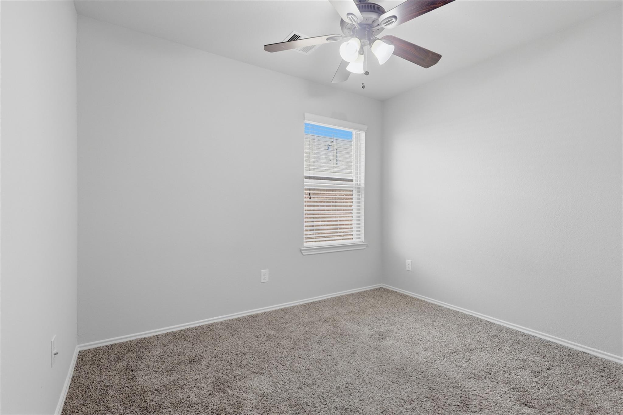 Empty bedroom with beige carpet, a dark-bladed ceiling fan, and a window with white blinds.