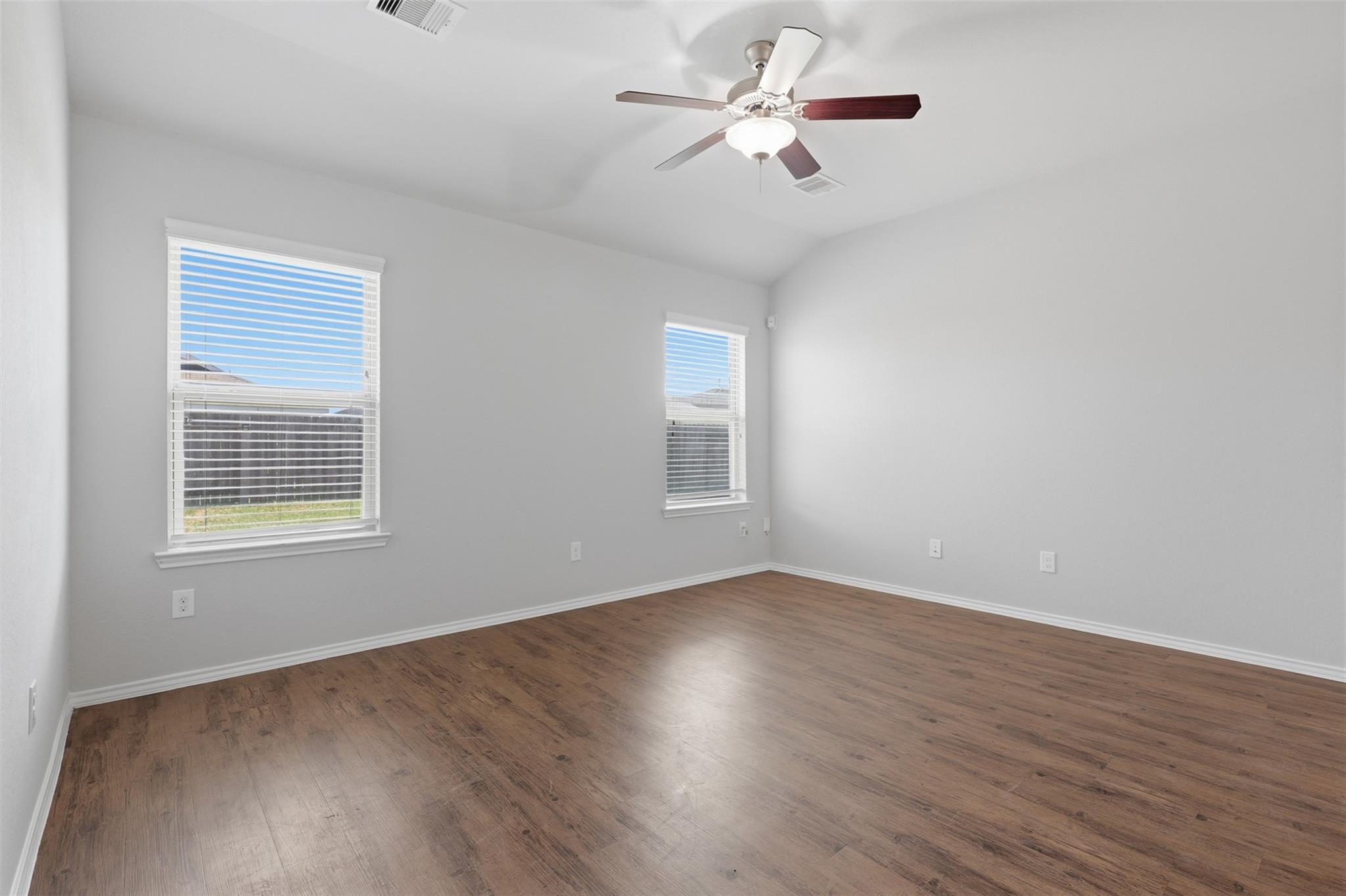 Empty room with brown wood-look flooring, two windows with blinds, and a ceiling fan.