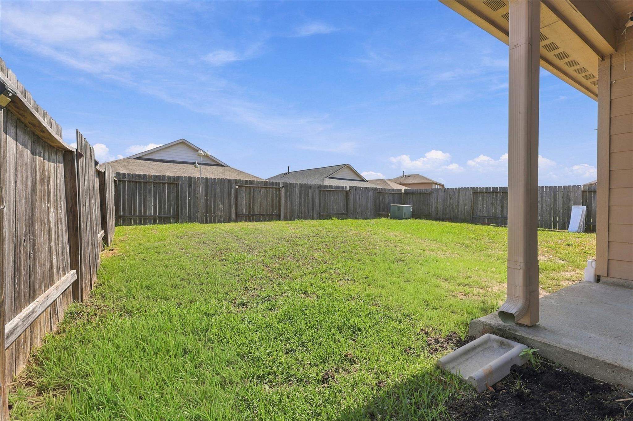 Fenced grassy backyard viewed from a covered patio, with neighboring homes beyond the fence.