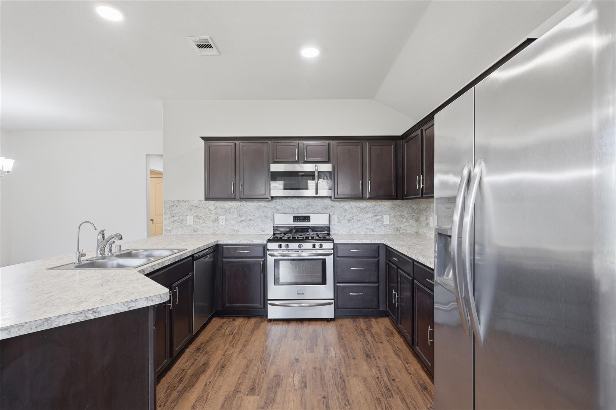 Kitchen with dark cabinets, stainless steel appliances, light stone counters, and wood-look flooring.