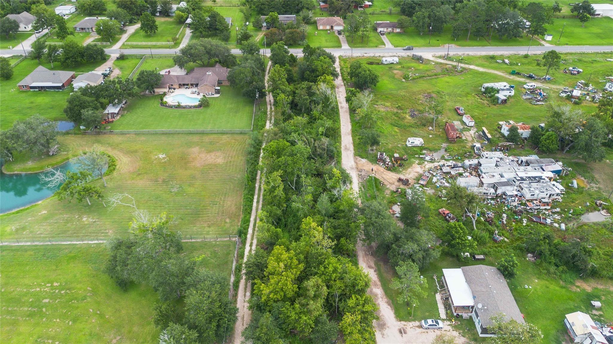 Aerial view of a grassy property with a pond, tree-lined driveway, and neighboring homes.
