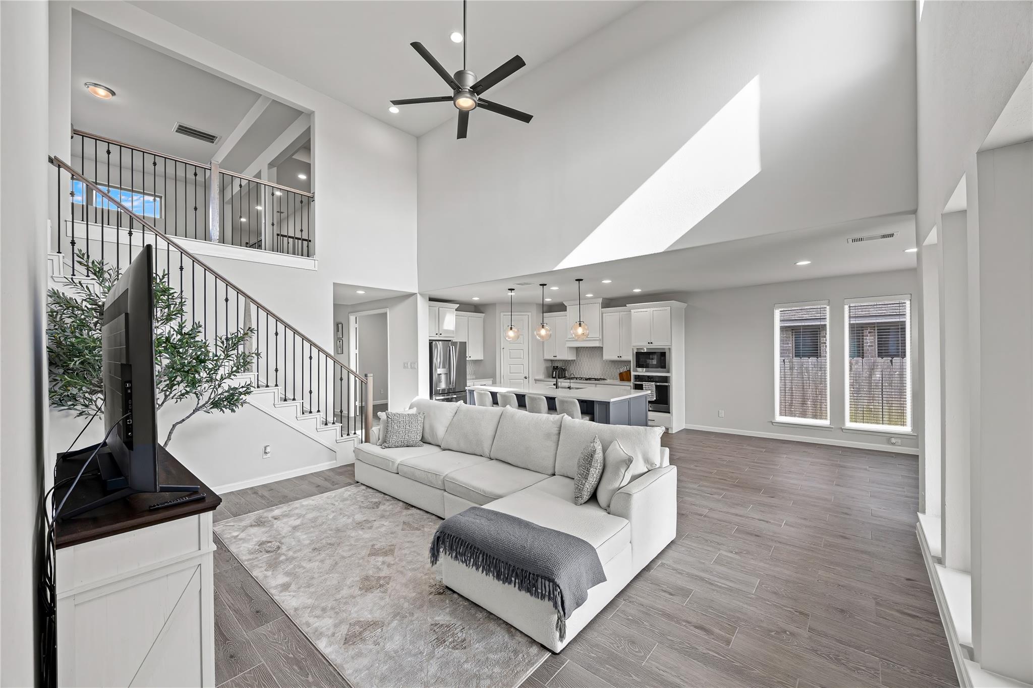 Open-plan living room with white sectional, staircase, and kitchen beneath a high ceiling.