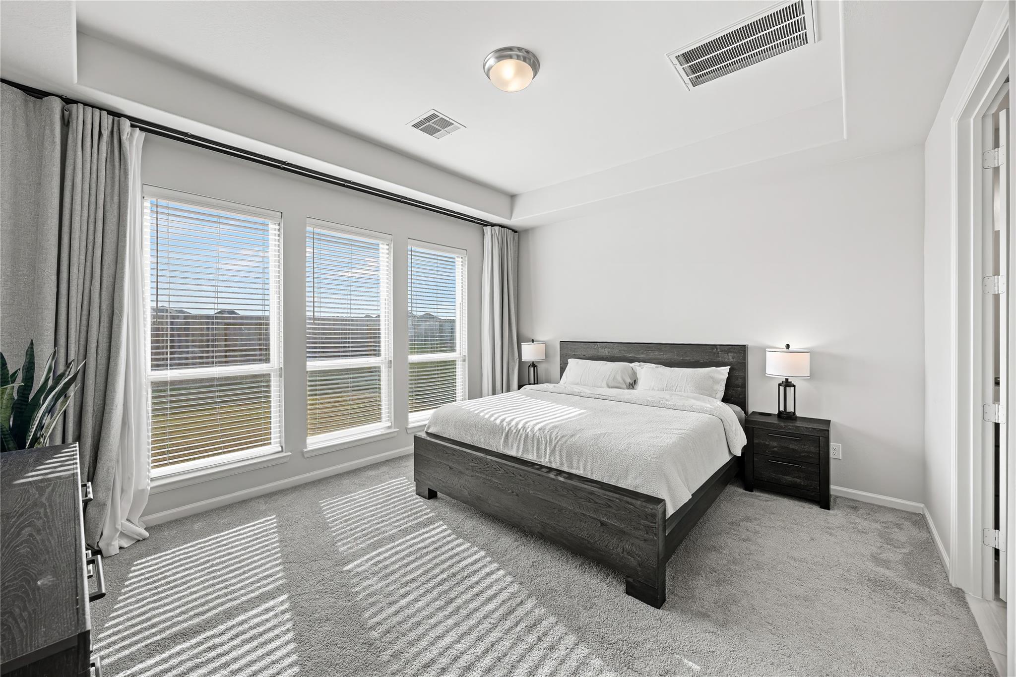 Bedroom with a dark wood bed and three curtained windows casting striped sunlight across the gray carpet.