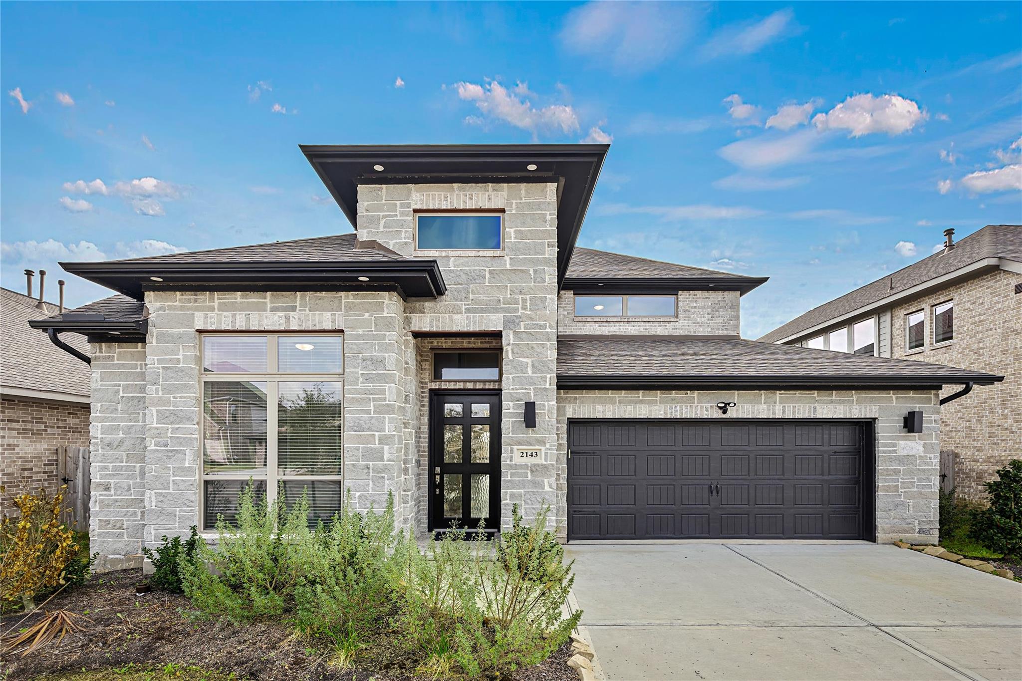 Gray stone two-story home with a black double garage door, wide driveway, and landscaped front shrubs.