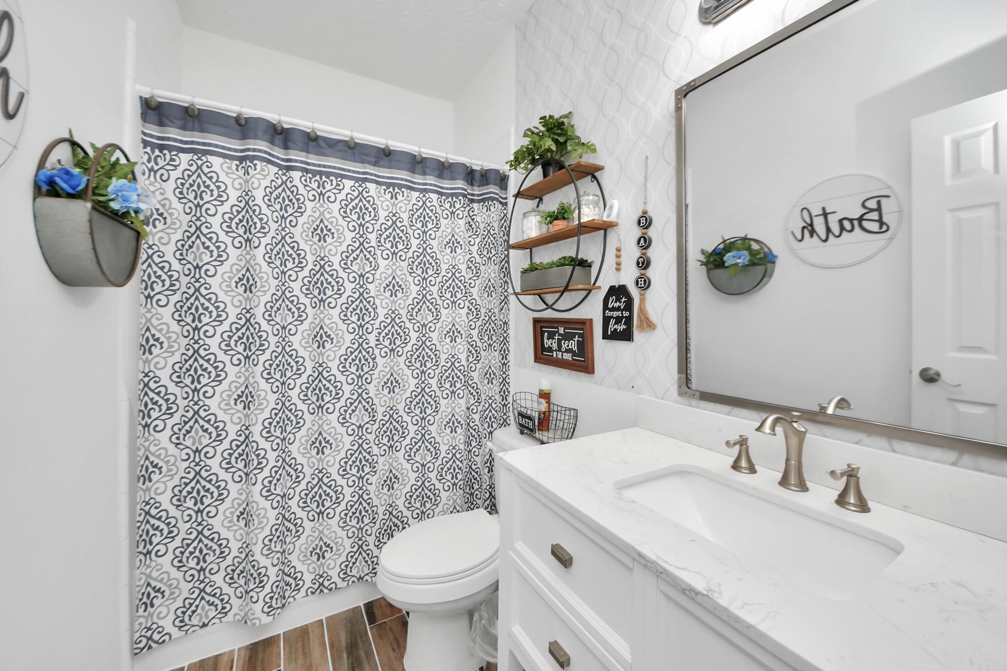 Bathroom with double-sink vanity, patterned shower curtain, toilet, and wood-look tile floor.
