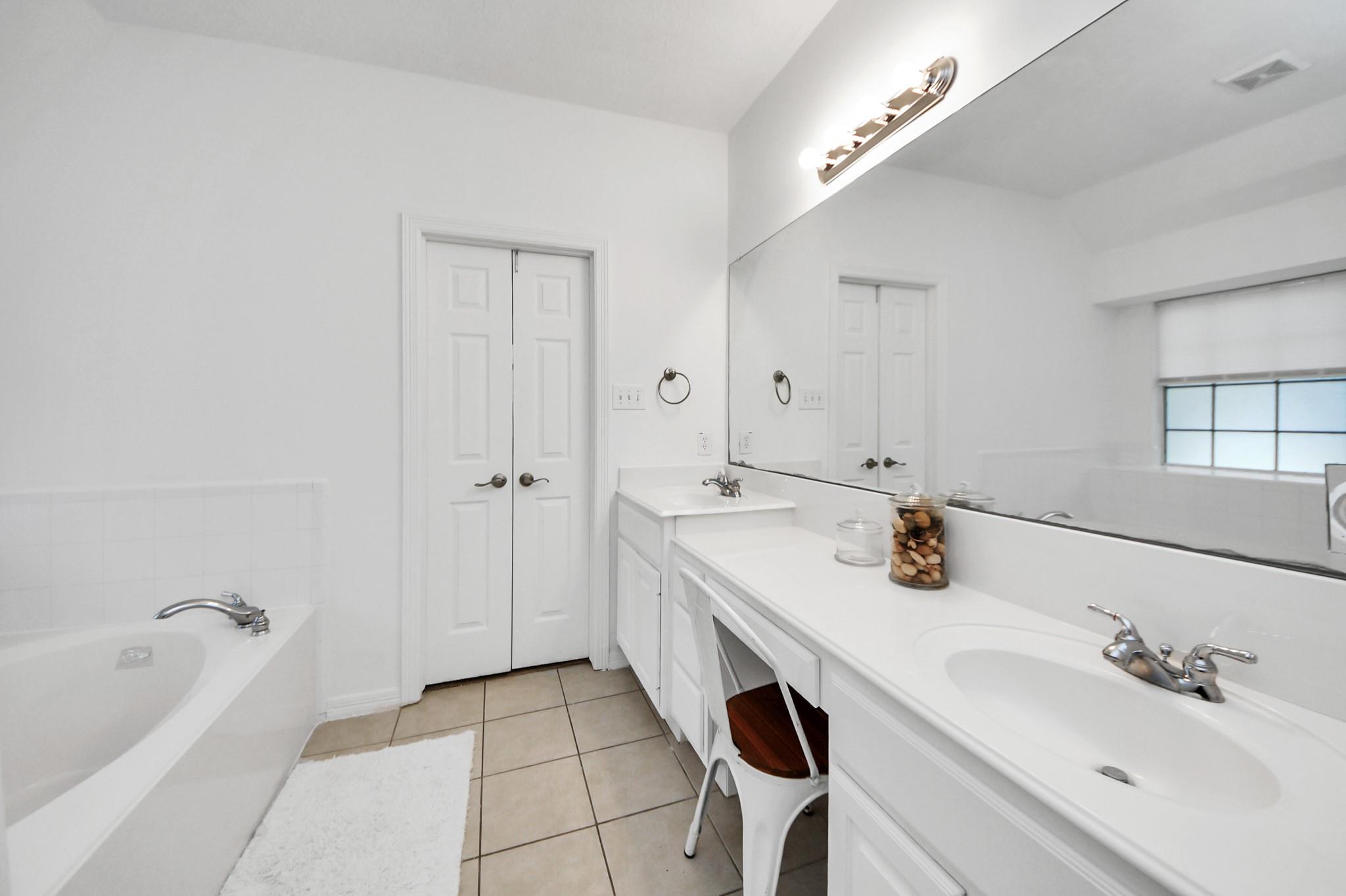 Bright bathroom with a bathtub, double-sink vanity, large mirror, and beige tiled floor.