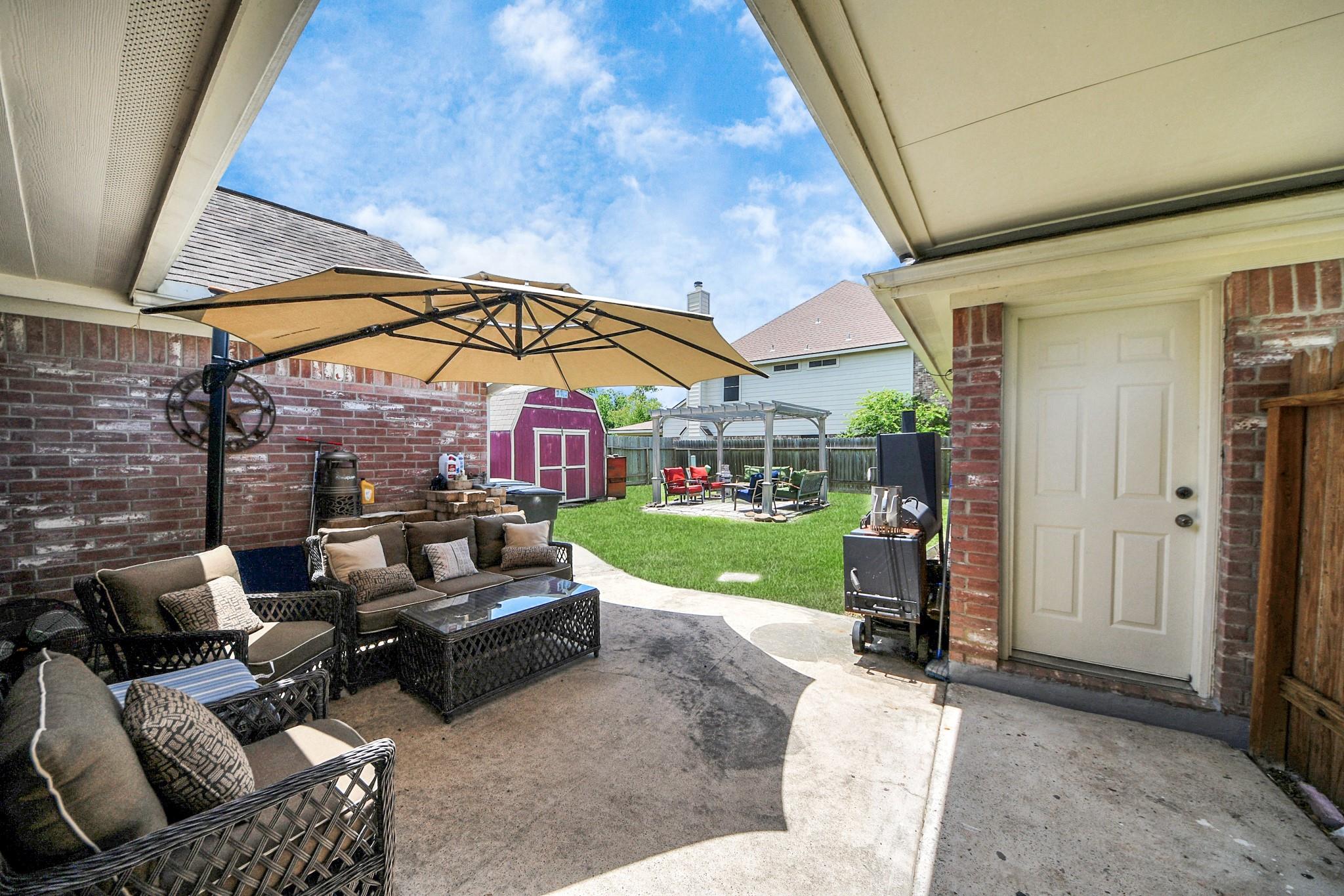 Covered patio with wicker seating and a tan umbrella overlooks a lawn, red shed, and pergola.