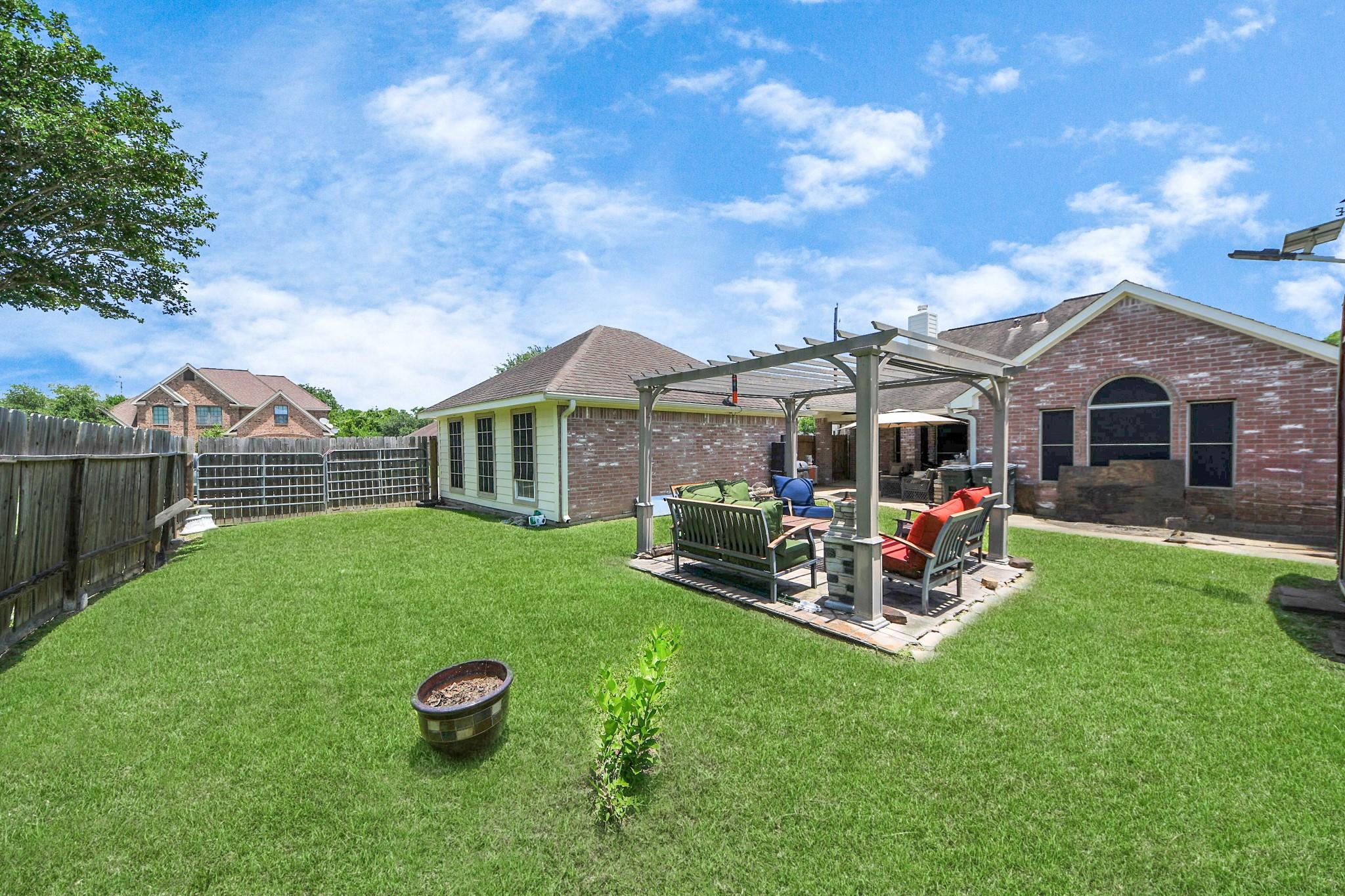 Fenced backyard with a furnished pergola patio beside a brick house and broad green lawn.