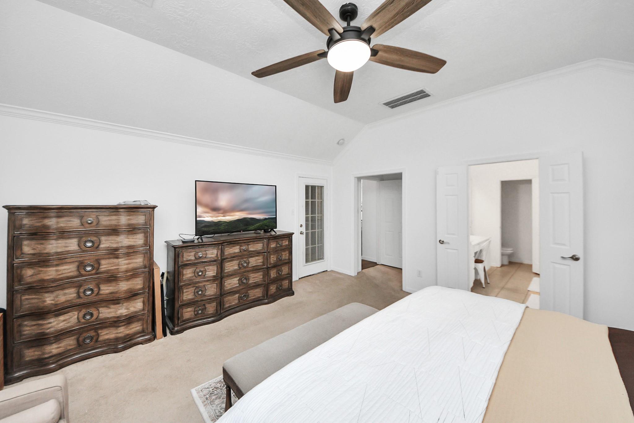Furnished bedroom with a vaulted ceiling, dark wood dressers, and open doors to an adjoining bathroom.