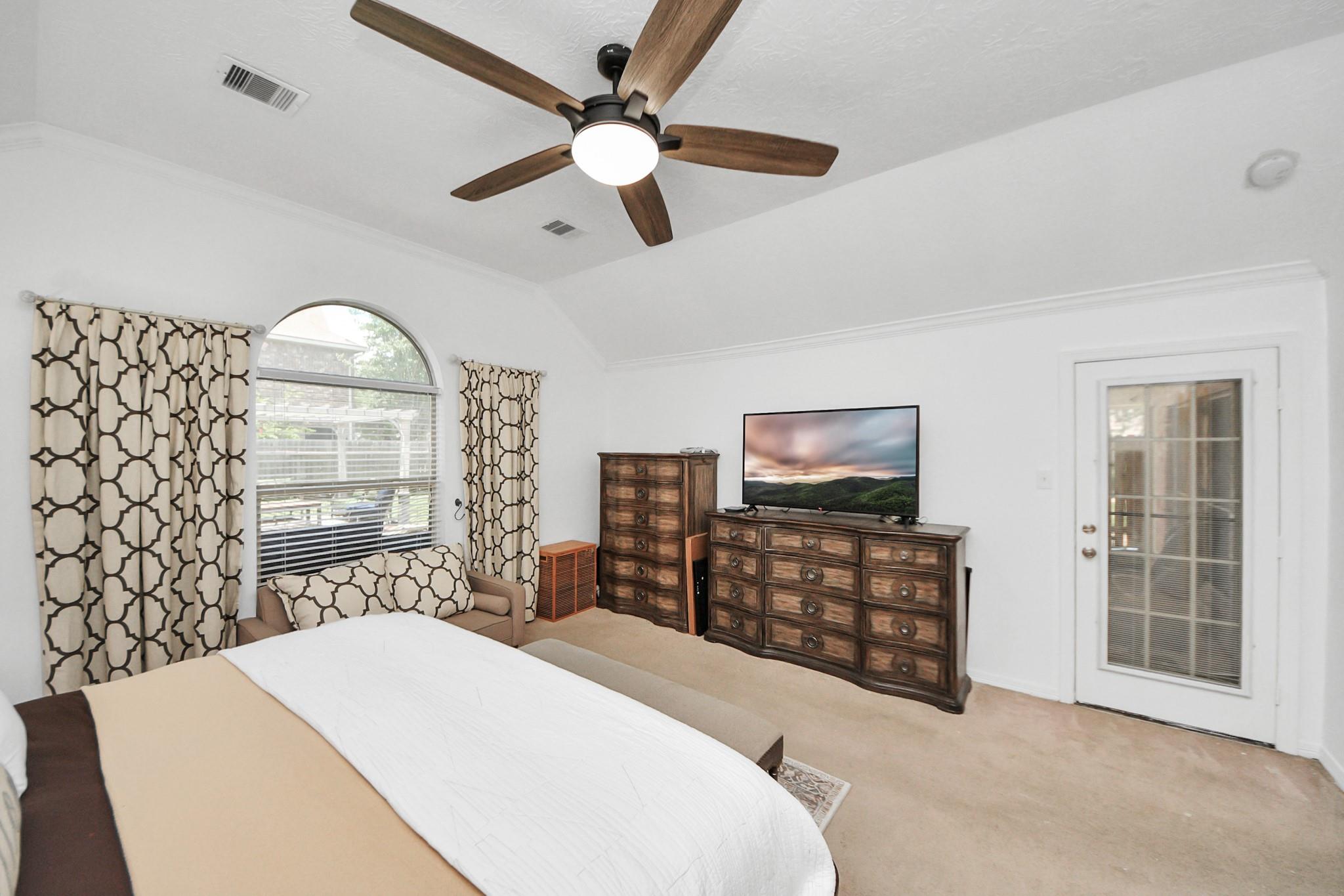 Furnished bedroom with an arched window, patterned curtains, dark wood dressers, and a ceiling fan.