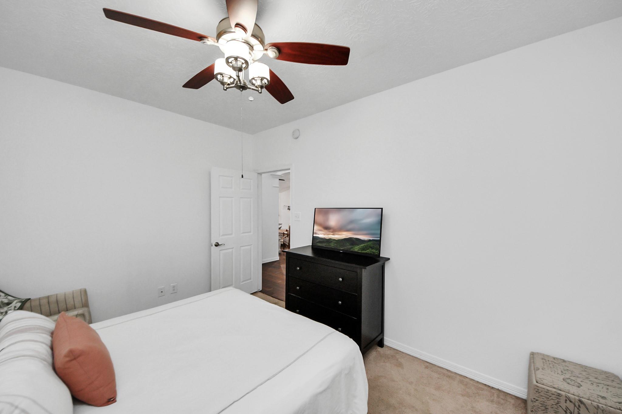 Bedroom with a white bed, dark dresser and television, and ceiling fan.