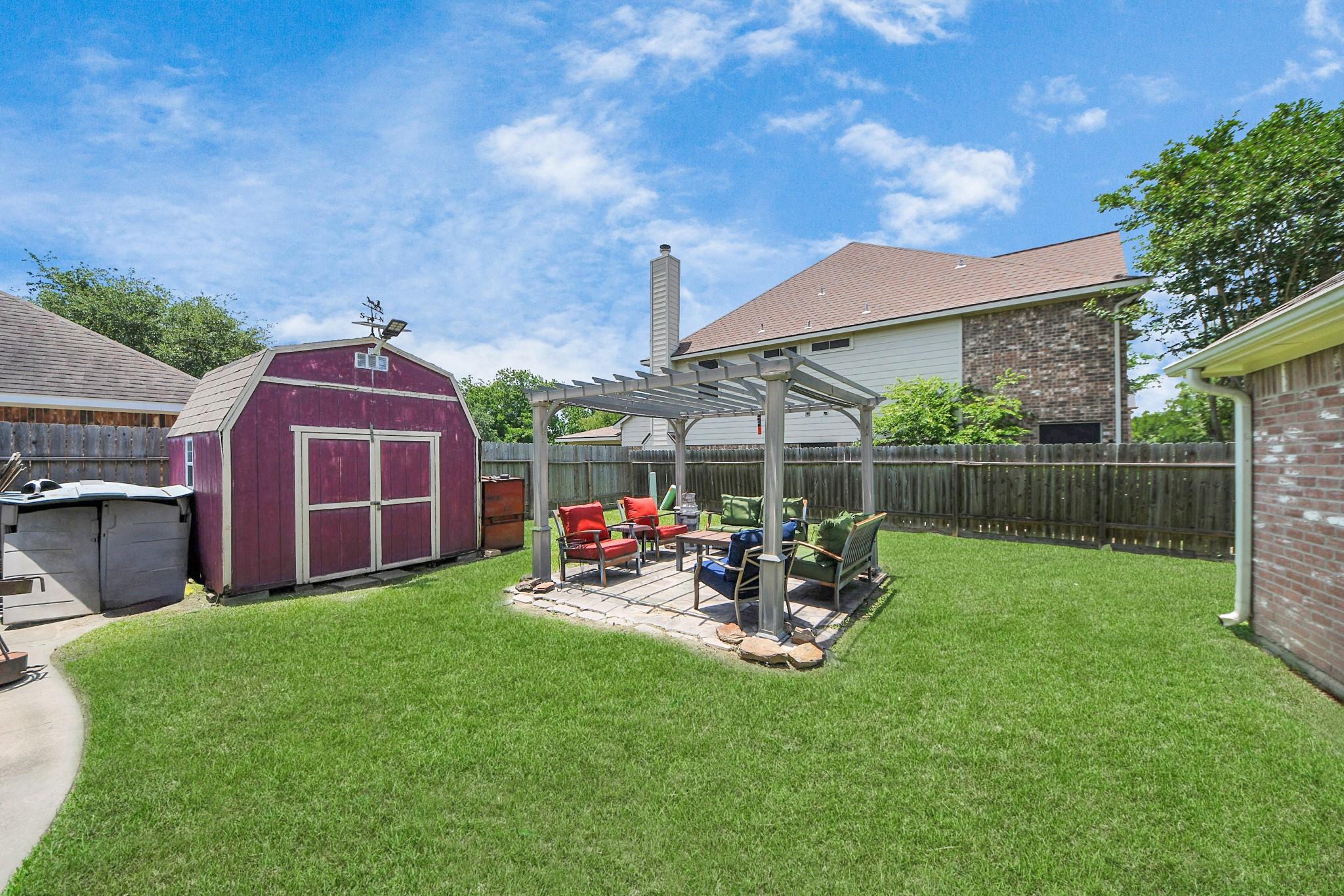 Backyard with a red shed and furnished patio beneath a pergola, surrounded by a fenced green lawn.