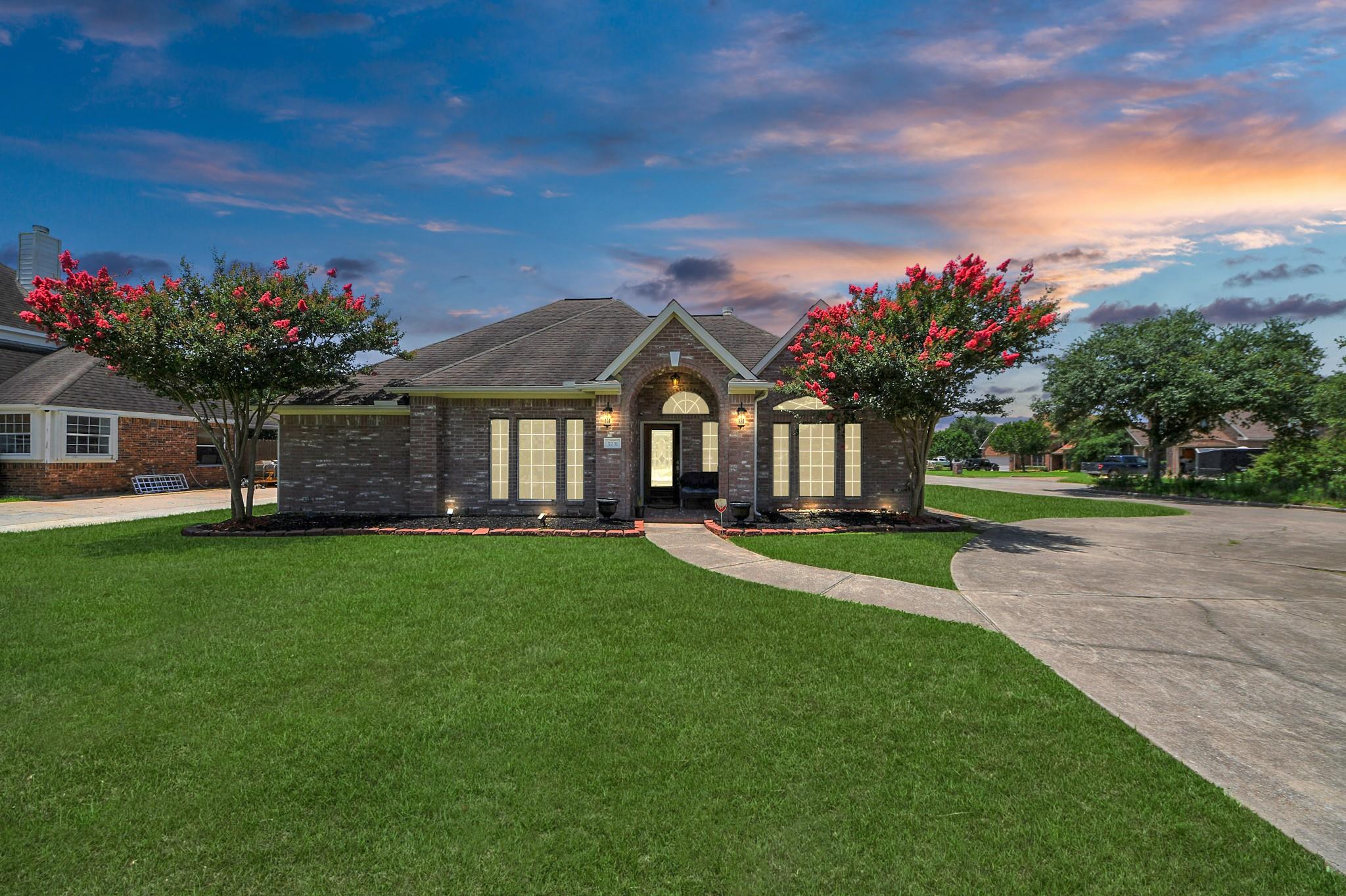 Brick house with a gabled entry, broad lawn, curved driveway, and flowering trees at sunset.